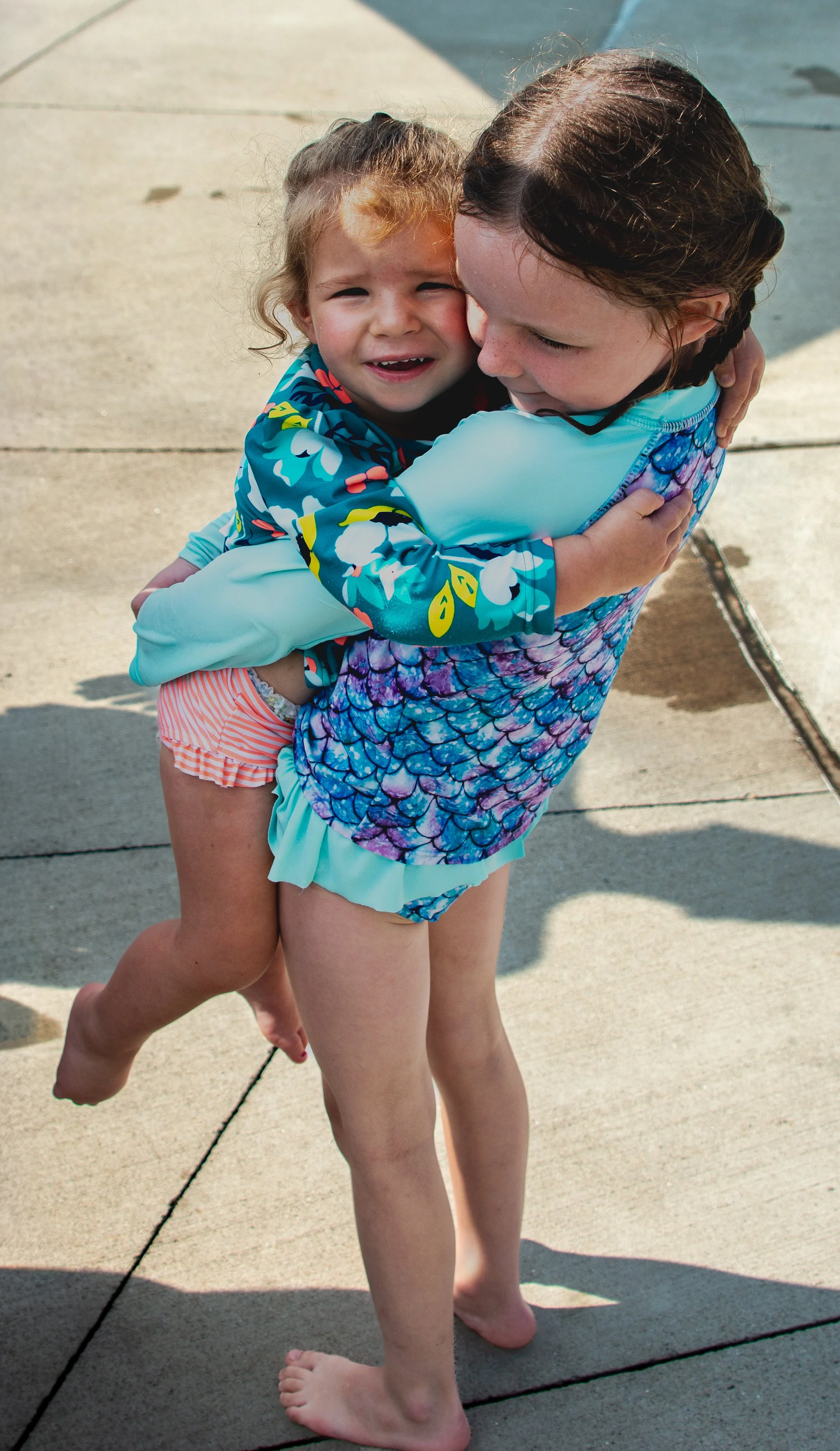 Two young girls hugging outdoors on a concrete surface, one girl is wearing a colorful swimsuit with a mermaid scale pattern and the other girl is wearing a long-sleeve floral top. They are barefoot.