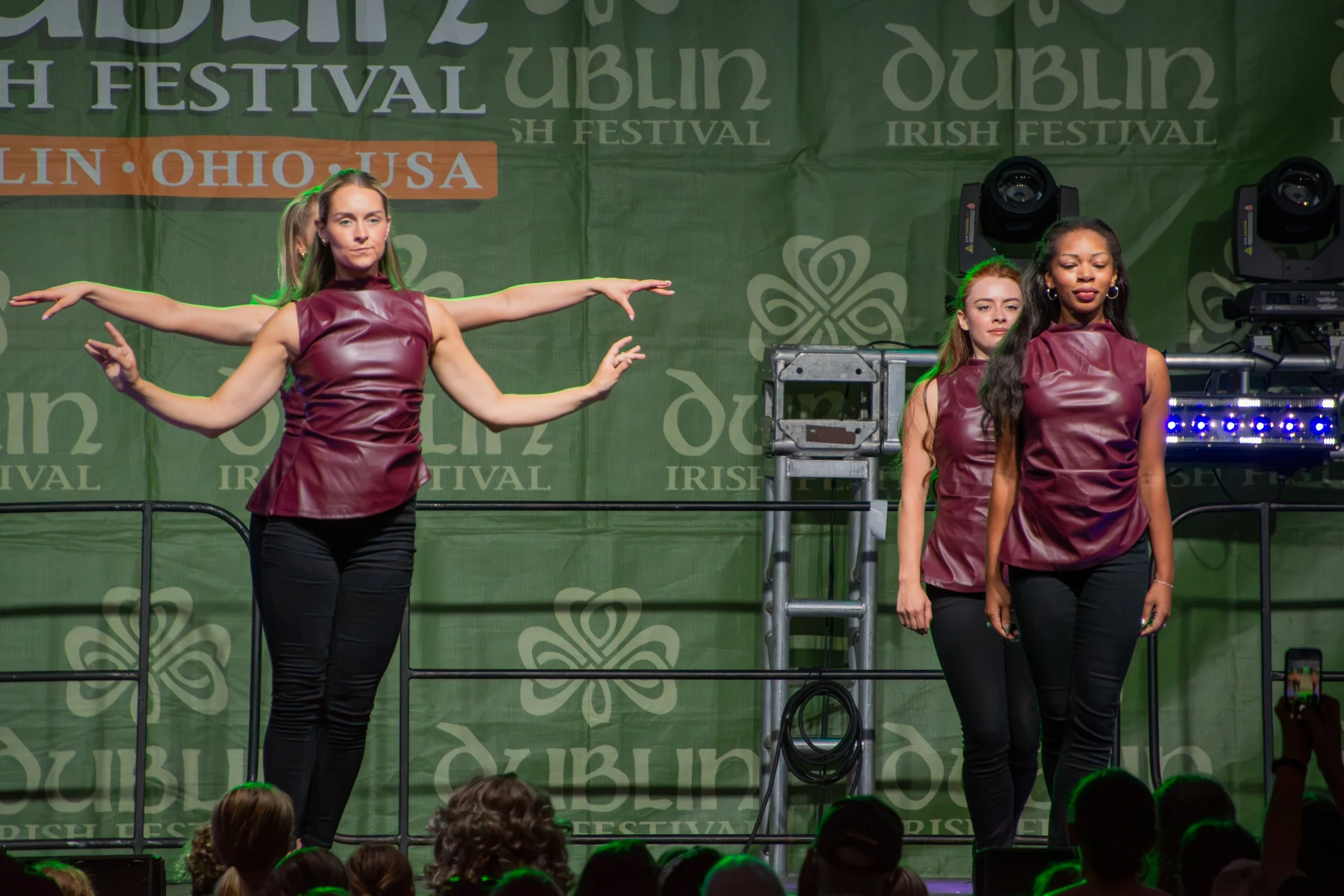 Three women performing Irish dance on stage at an Irish festival, with a green backdrop and audience in front.