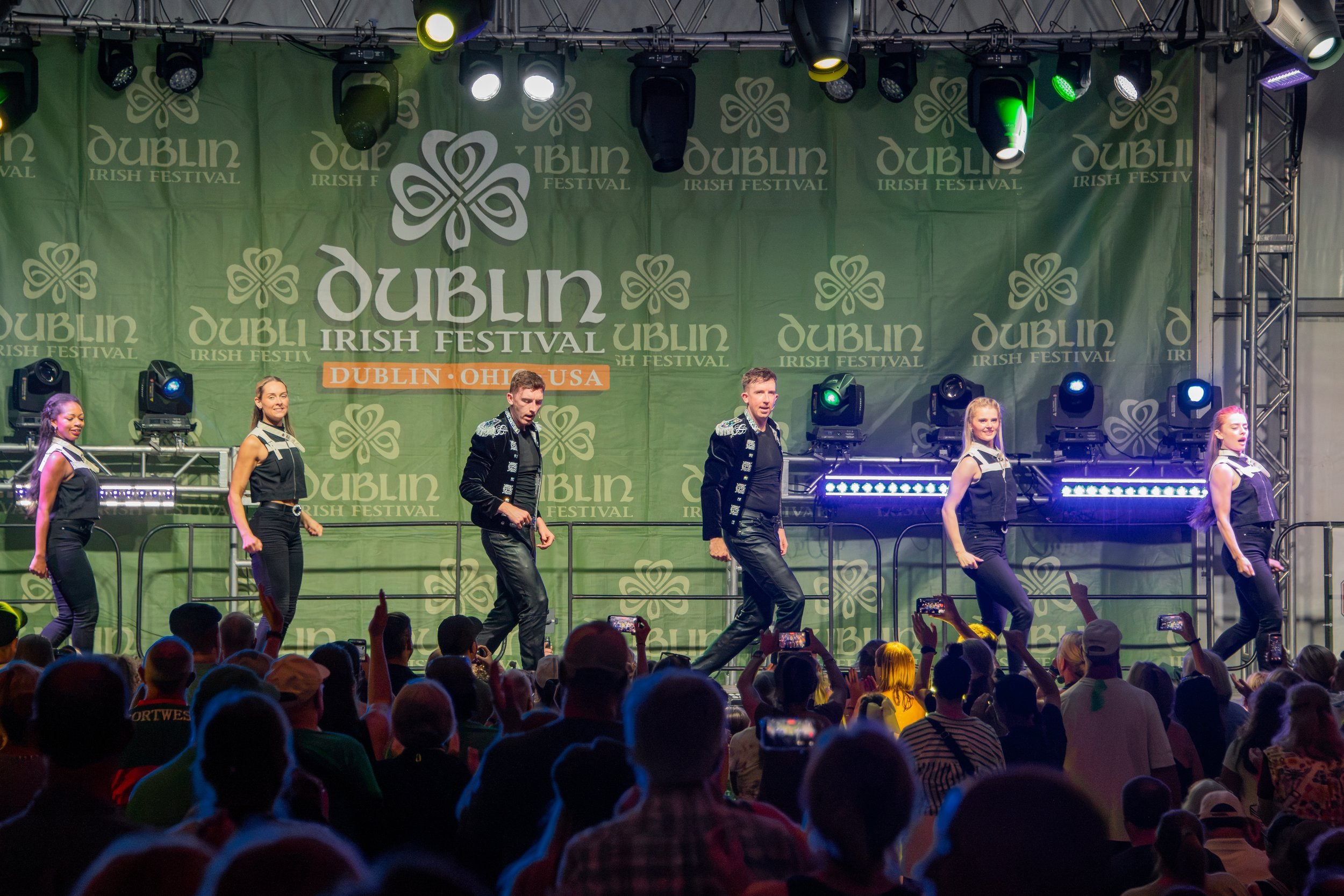 A stage at the Dublin Irish Festival with dancers performing in front of a large green backdrop displaying the festival's logo and name. Audience members are visible in the foreground, taking photos and watching the performance.