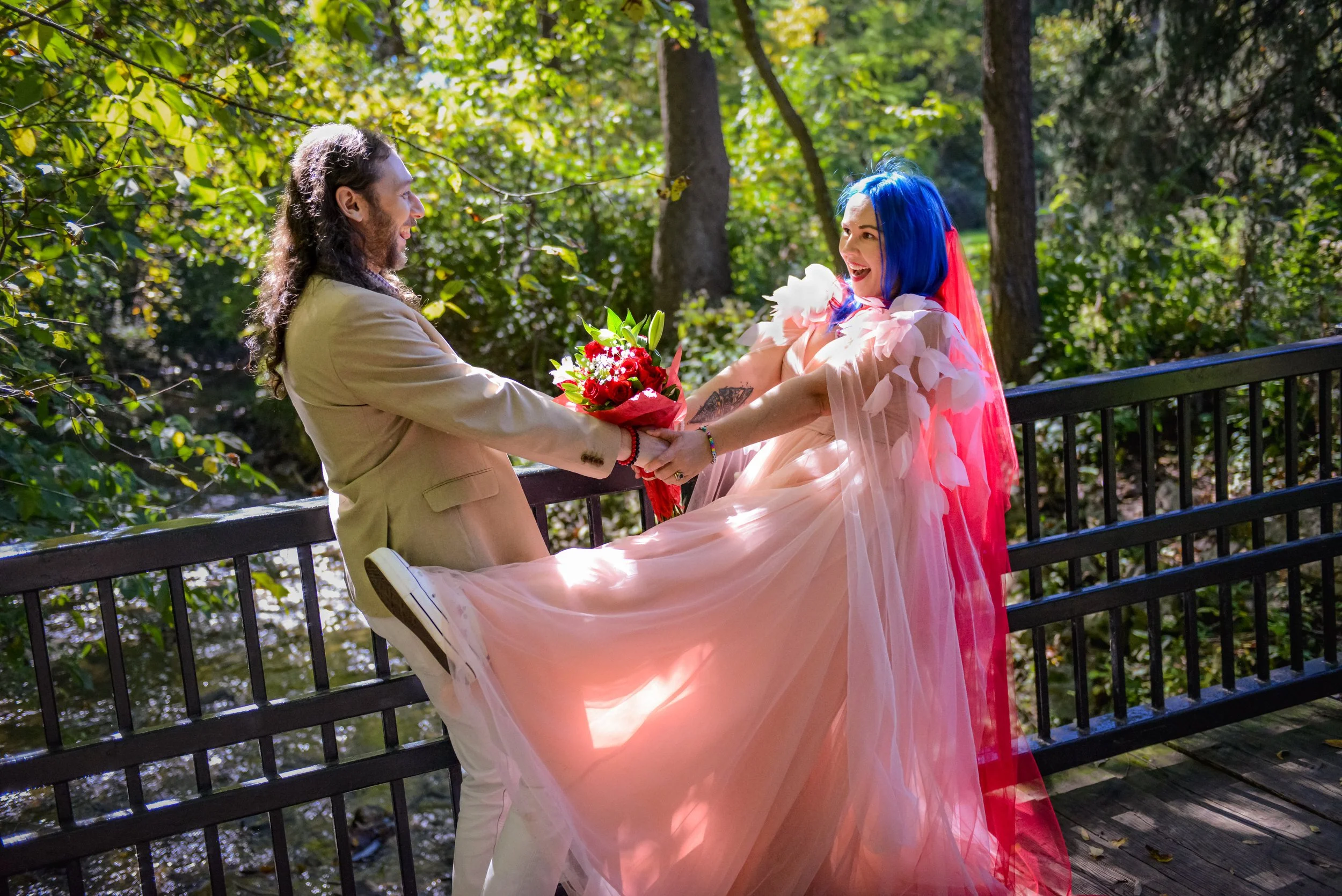 A person in a beige suit handing a bouquet of red and white flowers to a woman in a flowing pink dress with blue hair, sitting on a park bench surrounded by green trees.