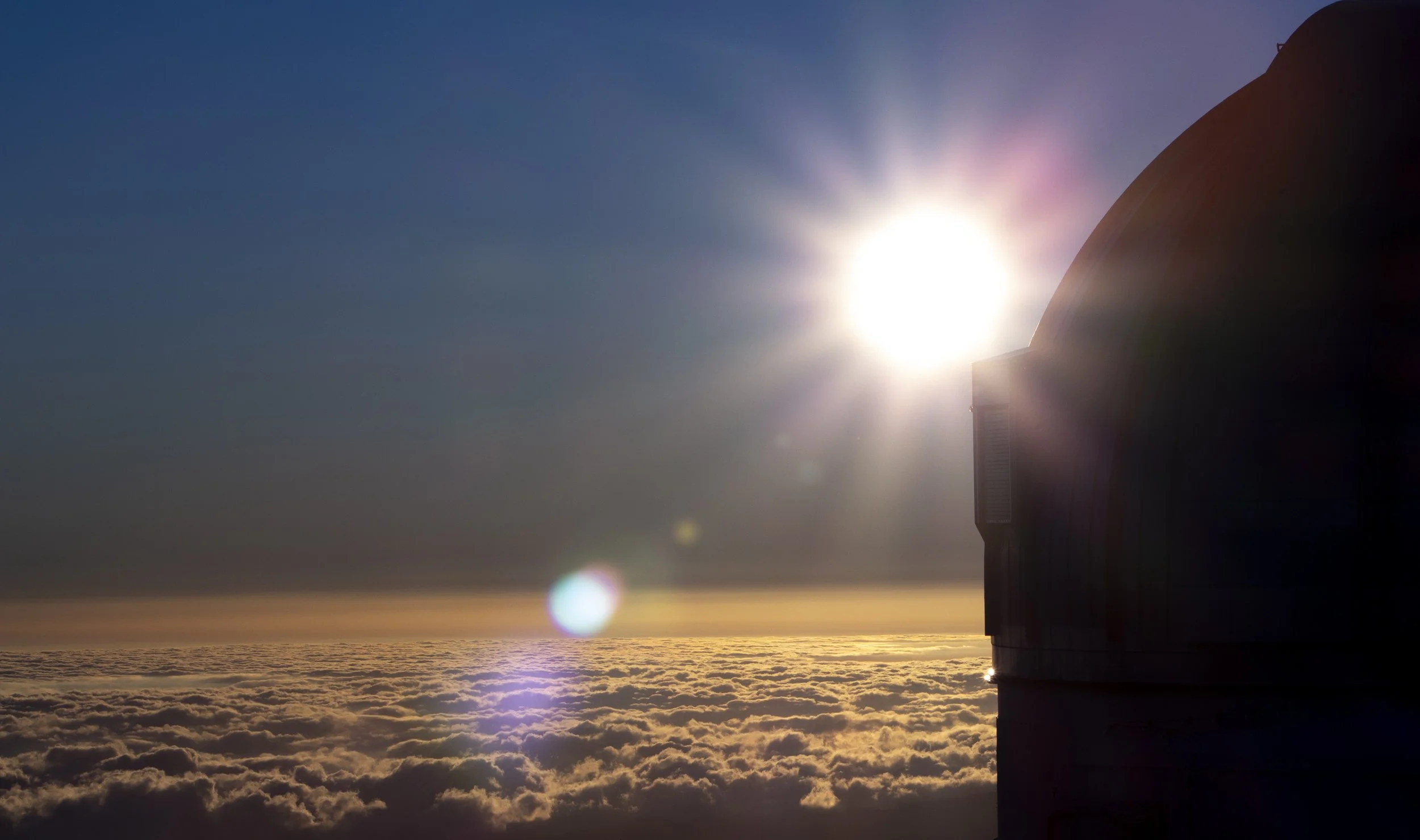 An aerial view of a building with a dome-shaped roof, partially silhouetted against a bright sun in the sky. The scene is above a thick layer of clouds during sunset or sunrise, with a clear horizon and lens flare effects.