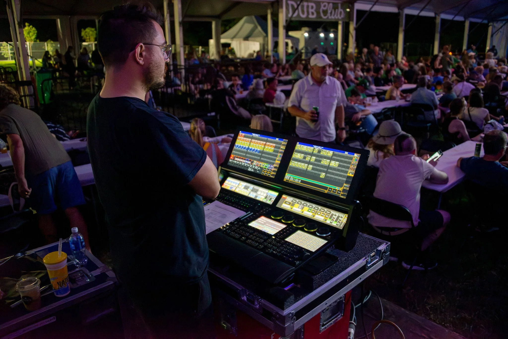 A DJ or music event performer stands in front of two digital mixing consoles with colorful displays, adjusting settings while a large crowd of people sits at tables under a covered outdoor venue at night, with a sign overhead that reads 'SUB Club'.