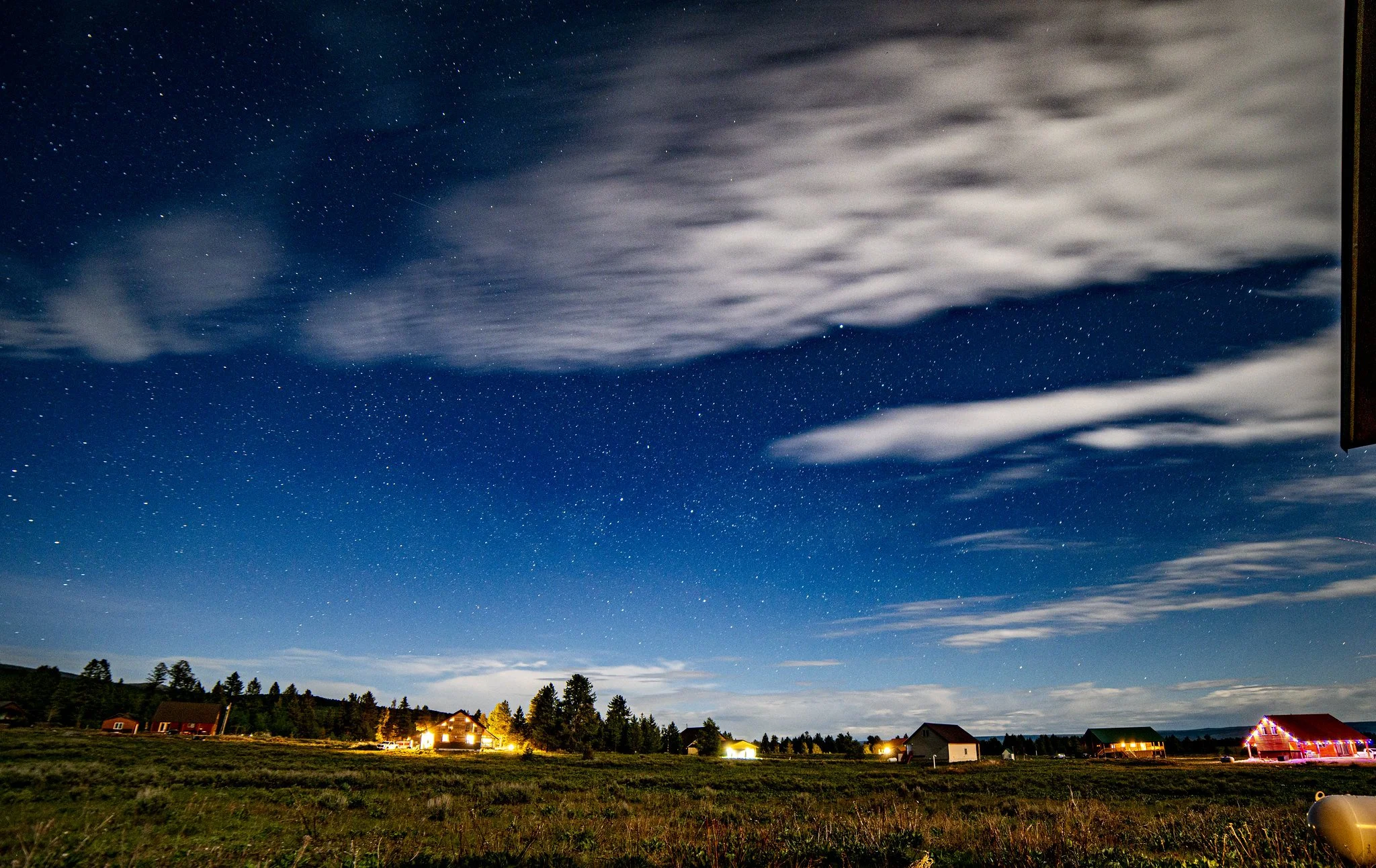 Nighttime scene of a rural area with houses, trees, and a cloudy sky. The sky is filled with stars and some clouds, with a dark landscape below.