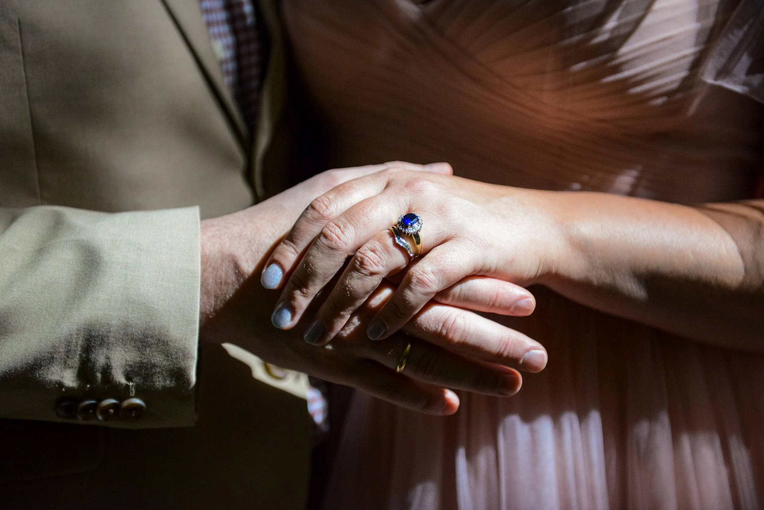 Close-up of two people's hands clasped together; one hand is wearing a ring with a blue gemstone.