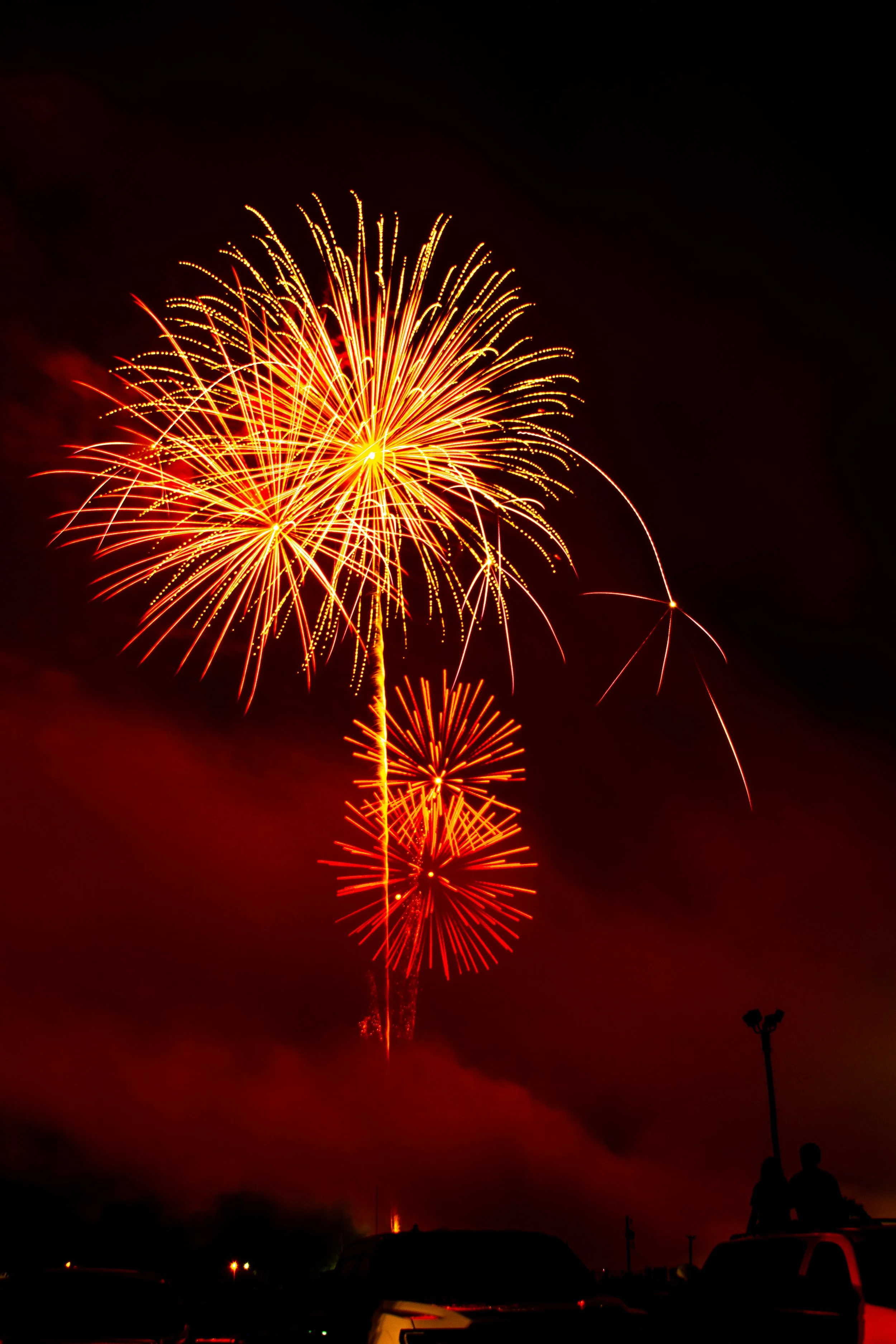 Colorful fireworks light up the dark night sky, with a reflection visible on the horizon and silhouetted people and vehicles in the foreground. Photographed by Cattywampus Camera at the Lancaster Ohio 4th of July fireworks show.