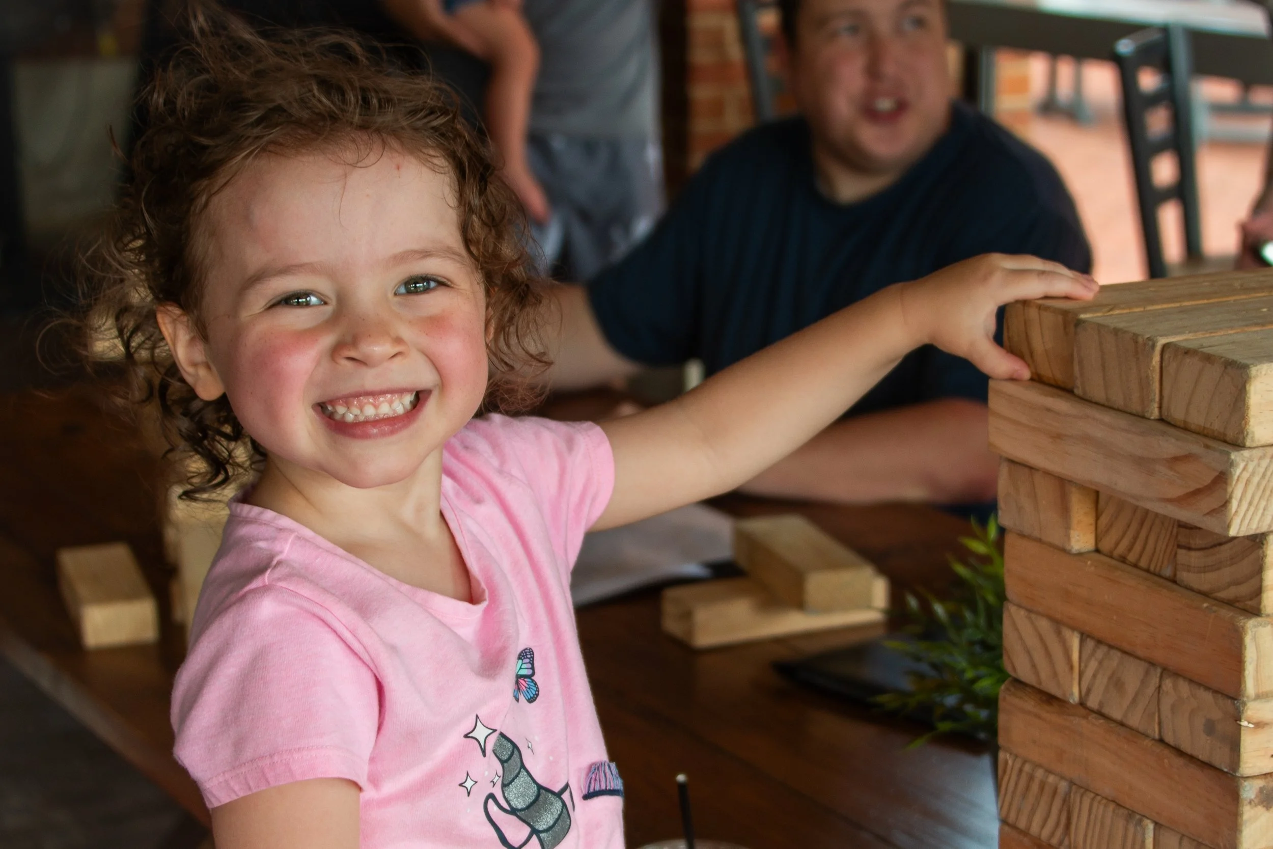 A young girl with curly hair and a pink t-shirt smiling as she plays Jenga with an adult man in a dark shirt. They are in an indoor setting with brick walls and wooden furniture.