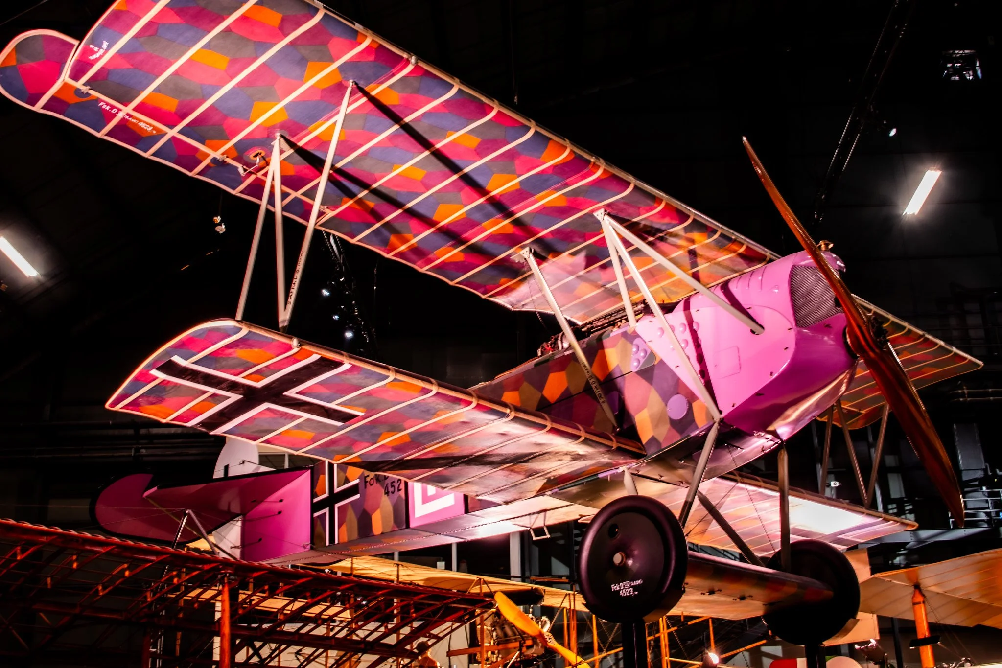 A colorful vintage biplane with pink and purple geometric patterned wings on display indoors, illuminated by bright lights.