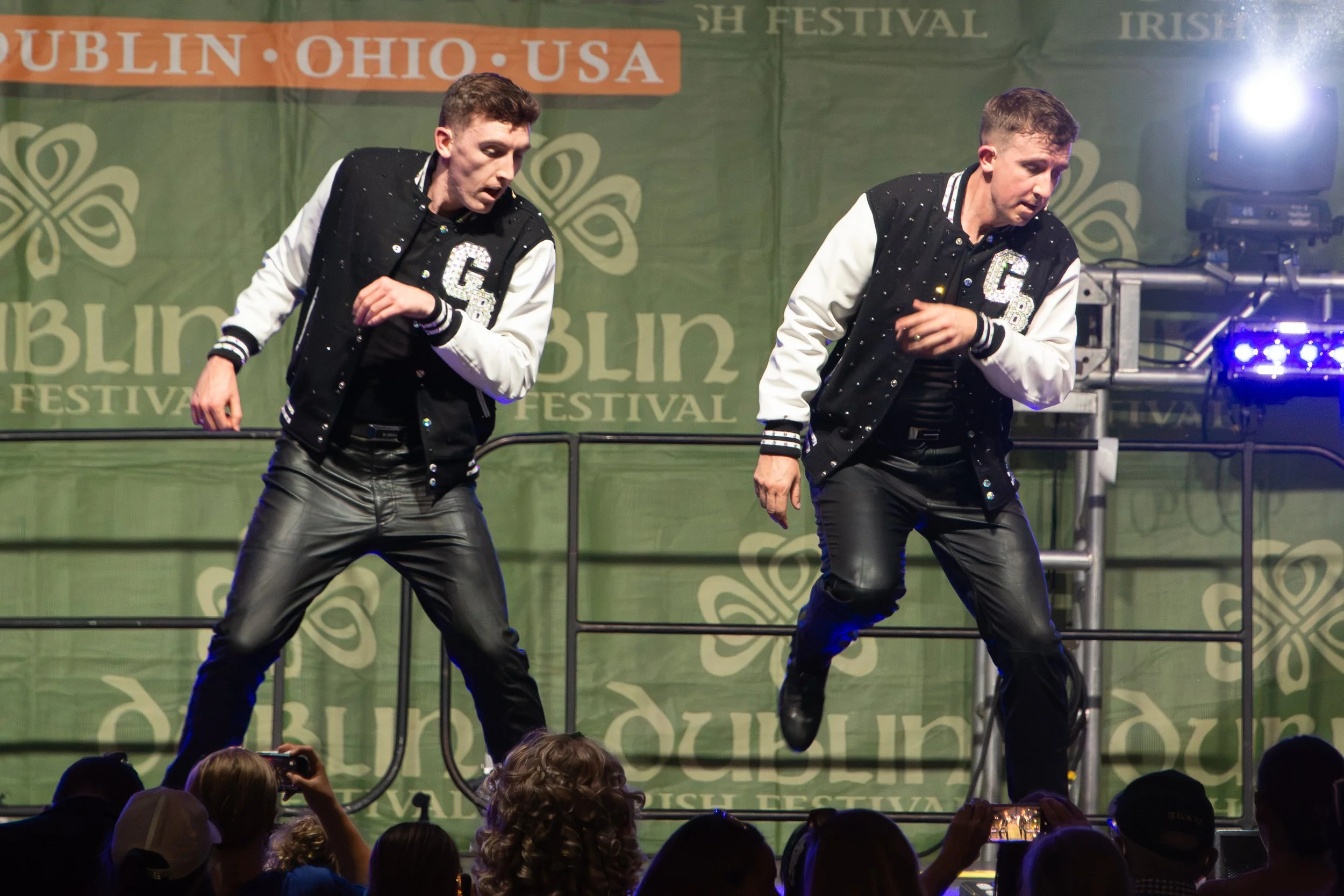 Two male performers dancing on stage during a St. Patrick's Day Festival, wearing black and white jackets with a letter 'C' and 'B' on them, with a green Festival backdrop and an audience watching.