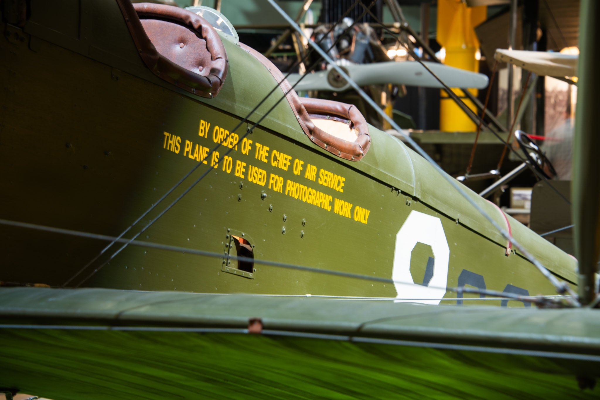 Close-up of a vintage biplane with a green body and brown leather seat, featuring yellow lettering indicating aviation regulations, with a blurred hangar or workshop in the background.