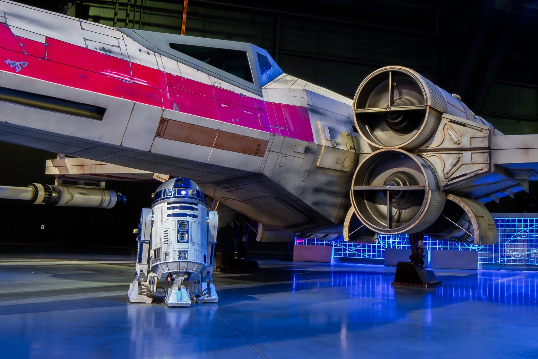 The image shows a detailed model of the Millennium Falcon from Star Wars, along with R2-D2 and BB-8 droids, displayed in a science fiction museum or exhibit with a blue grid background and neon lighting.