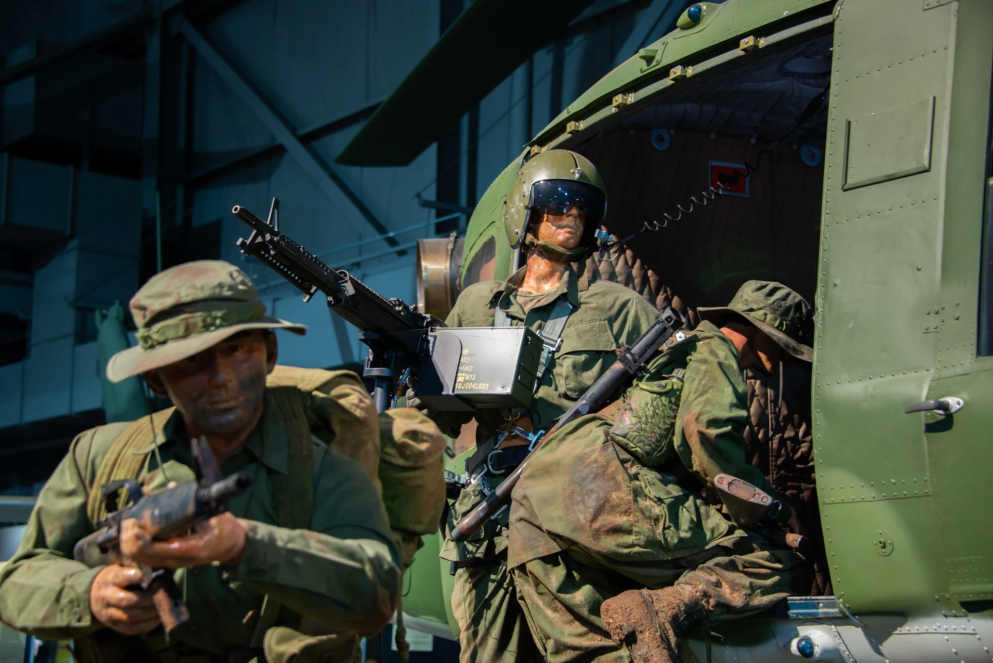 Military personnel in camouflage uniforms and helmets disembarking from a green helicopter, with a man holding a firearm in the foreground.