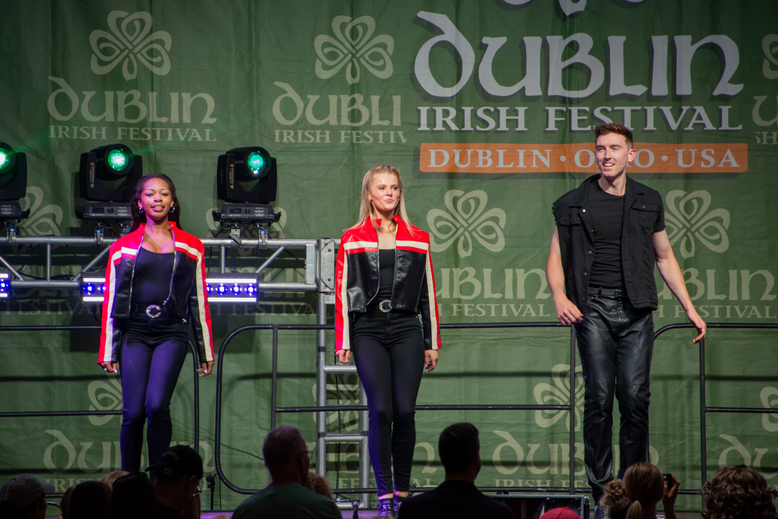 Three people, two women and one man, on stage at the Dublin Irish Festival in front of a green banner with festival logos and text, illuminated by stage lights, with audience members visible in the foreground.