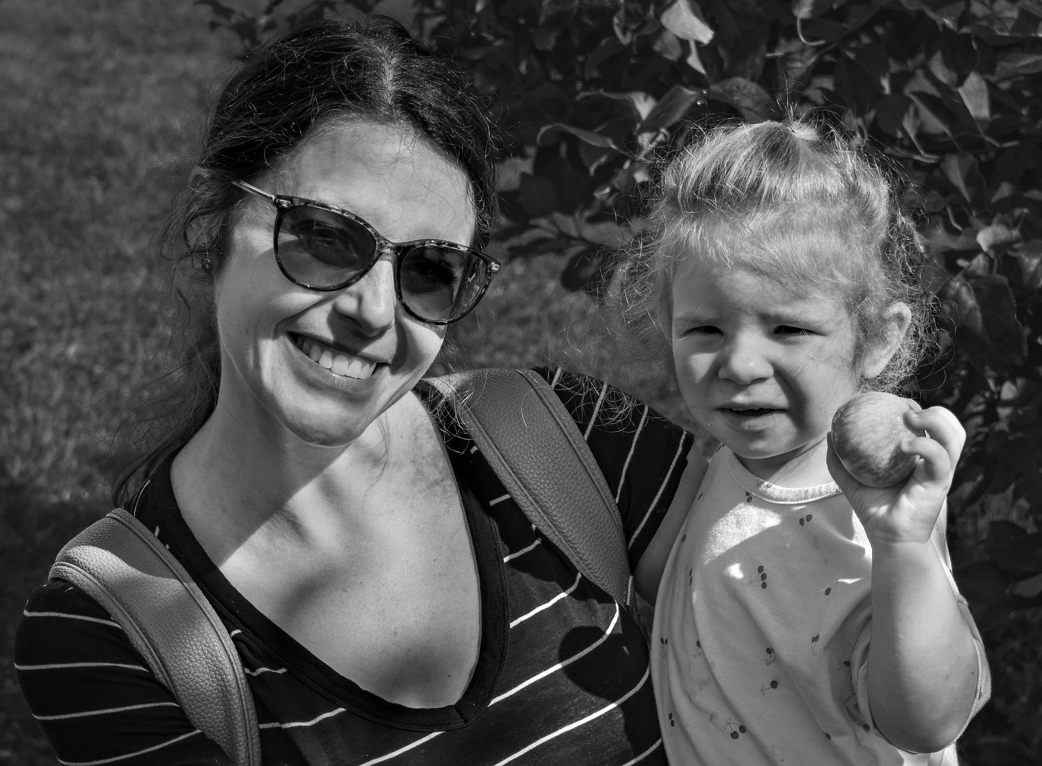 A woman wearing sunglasses smiling next to a young girl holding an apple, outdoors with trees in the background.