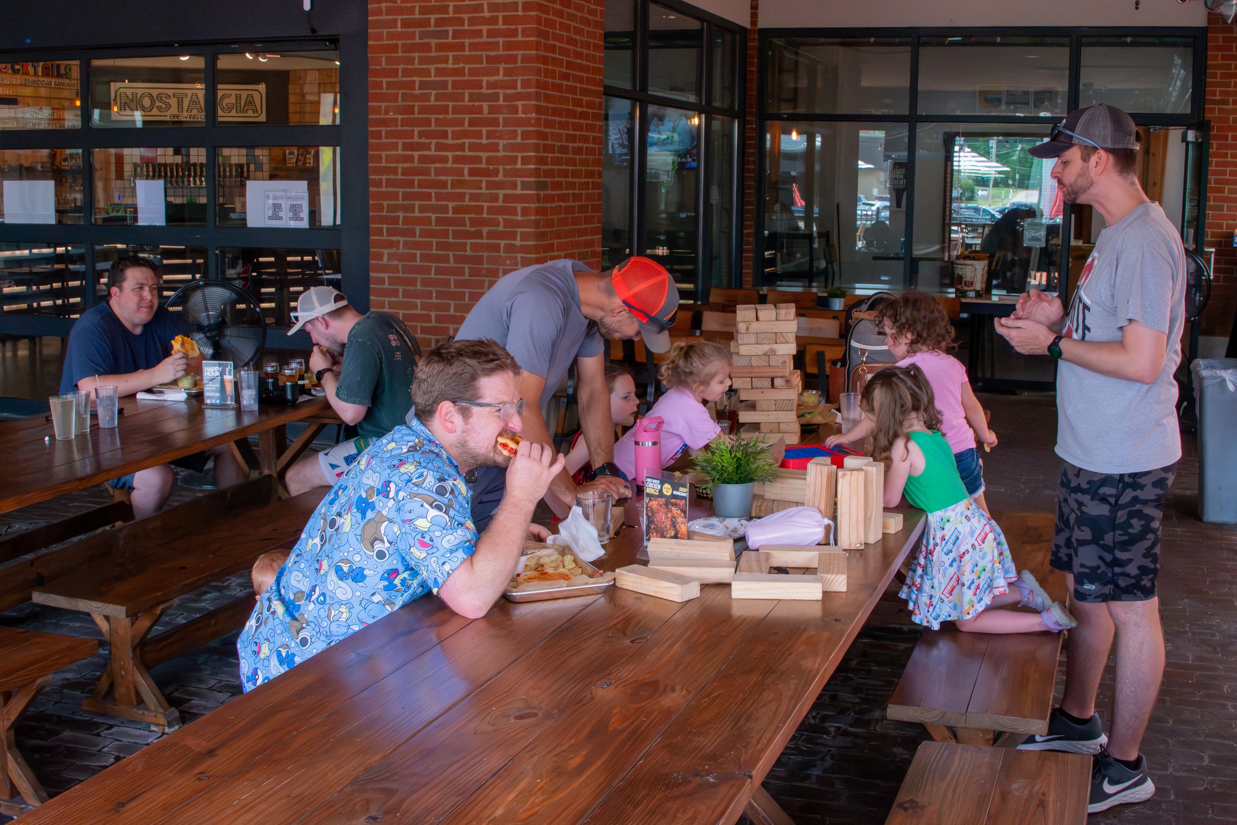 A diverse group of people, including children and adults, are gathered around a wooden table inside a restaurant. Some people are eating pizza, others are playing Jenga, and a man is standing on the right side, interacting with a child. The restauran