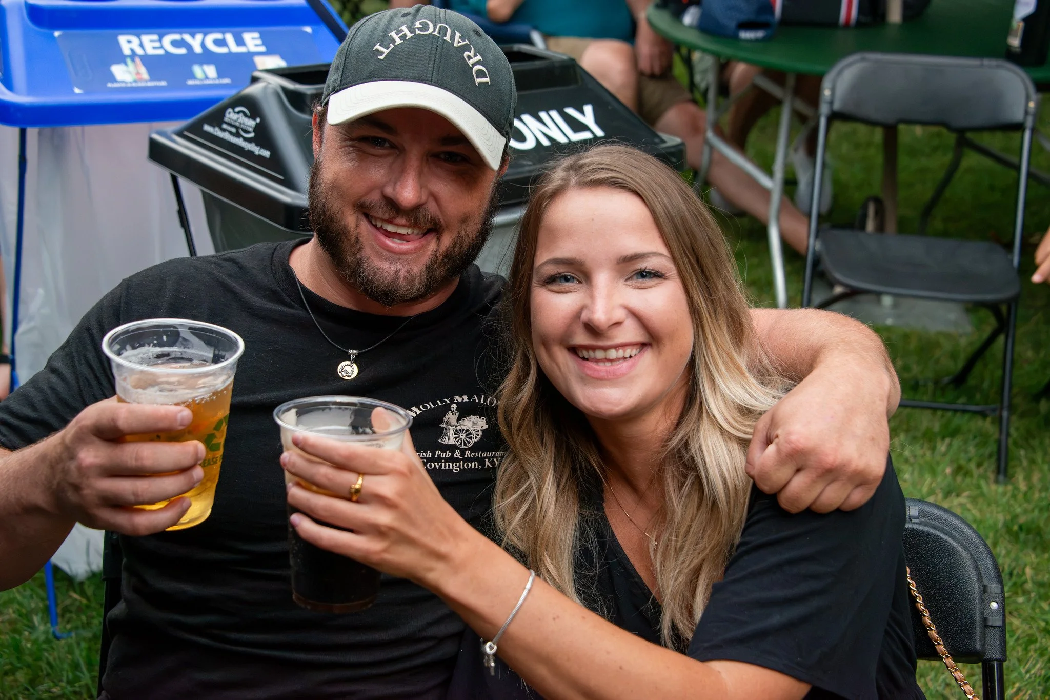 Two smiling adults, a man and a woman, are close together at an outdoor event, holding cups of beer, with people and chairs in the background.