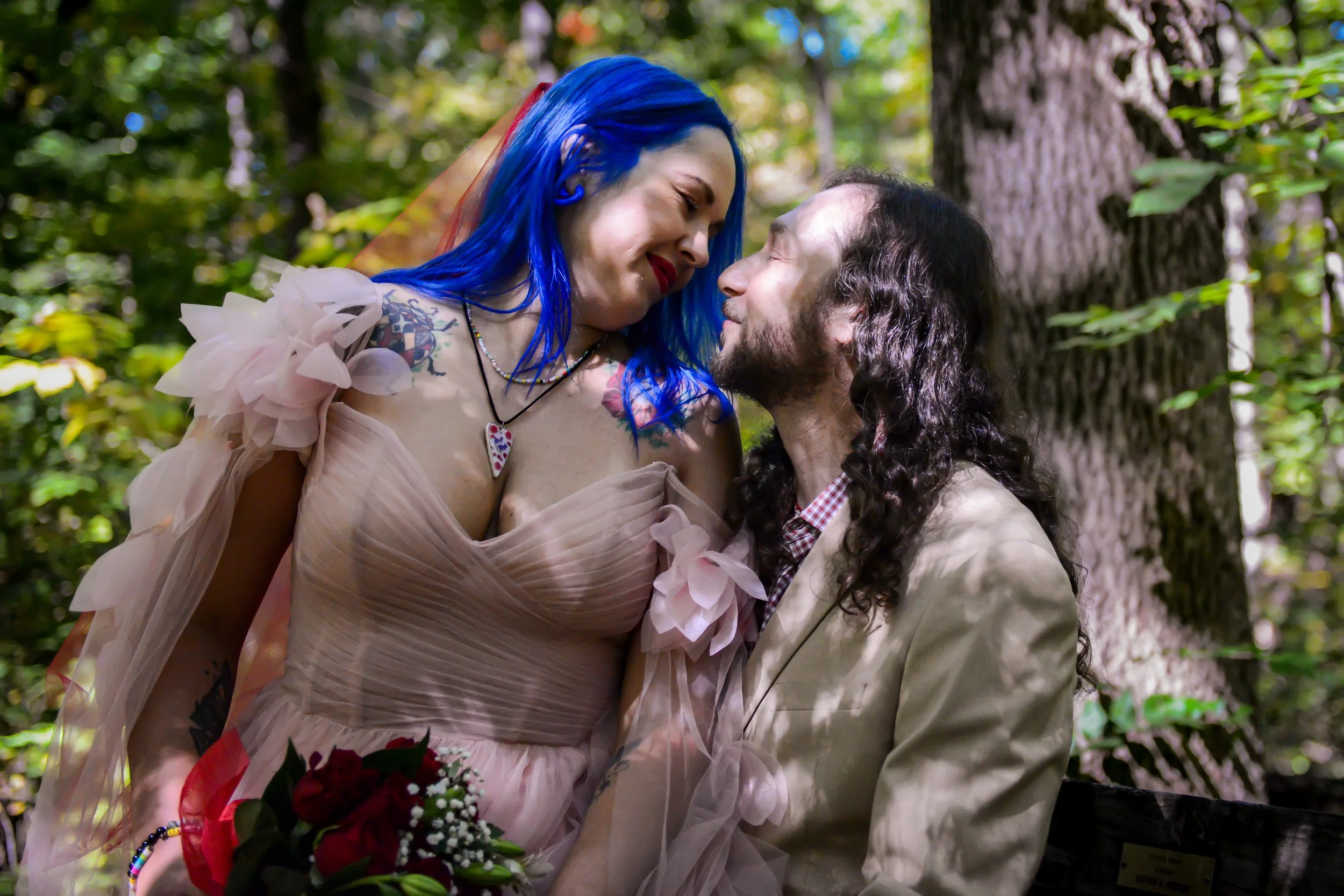 A couple is standing close together outdoors, with the woman smiling and the man looking at her. The woman has blue hair, is wearing a pink dress with ruffled sleeves, has colorful tattoos, and is holding a bouquet of red roses. The man has long dark curly hair, is dressed in a beige blazer and a checkered shirt, and is looking lovingly at her. They are surrounded by trees and greenery in a forest-like setting.