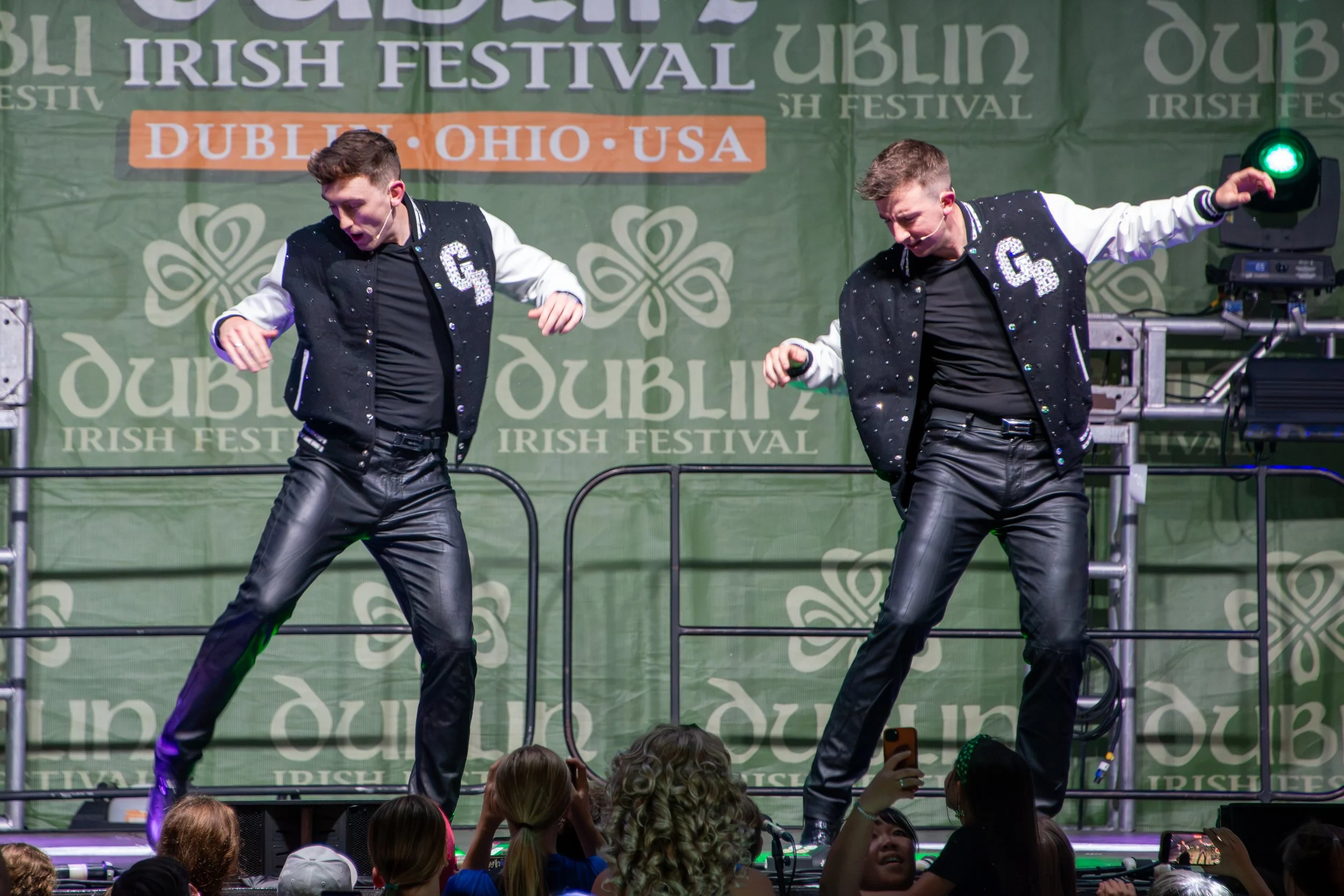 Two dancers performing on stage at an Irish festival, wearing black and white outfits with sequined jackets, in front of a green backdrop with festival signage.