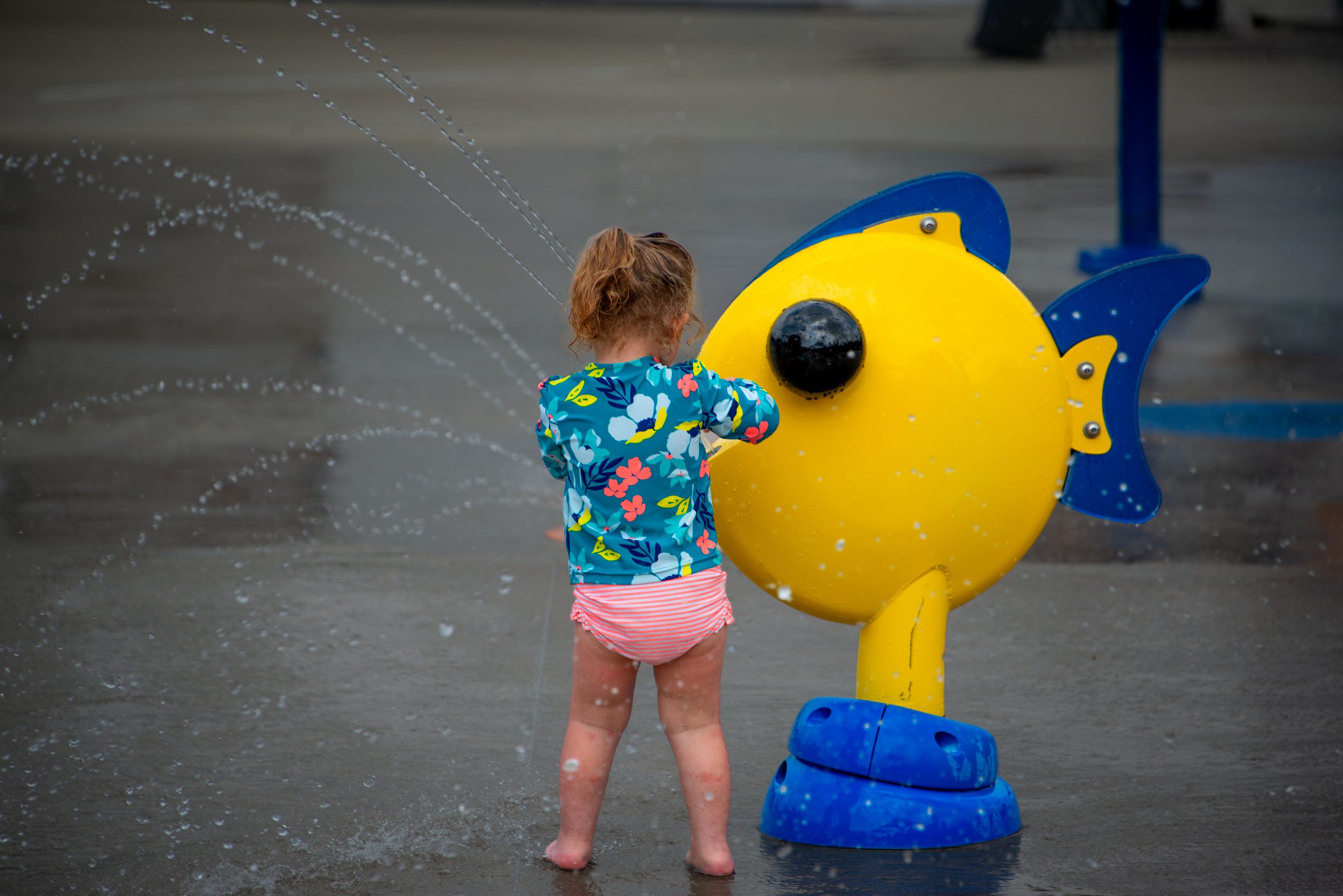 A young girl with curly hair, wearing a colorful jacket and pink striped shorts, playing in water at a playground with a yellow and blue fish-shaped water feature.