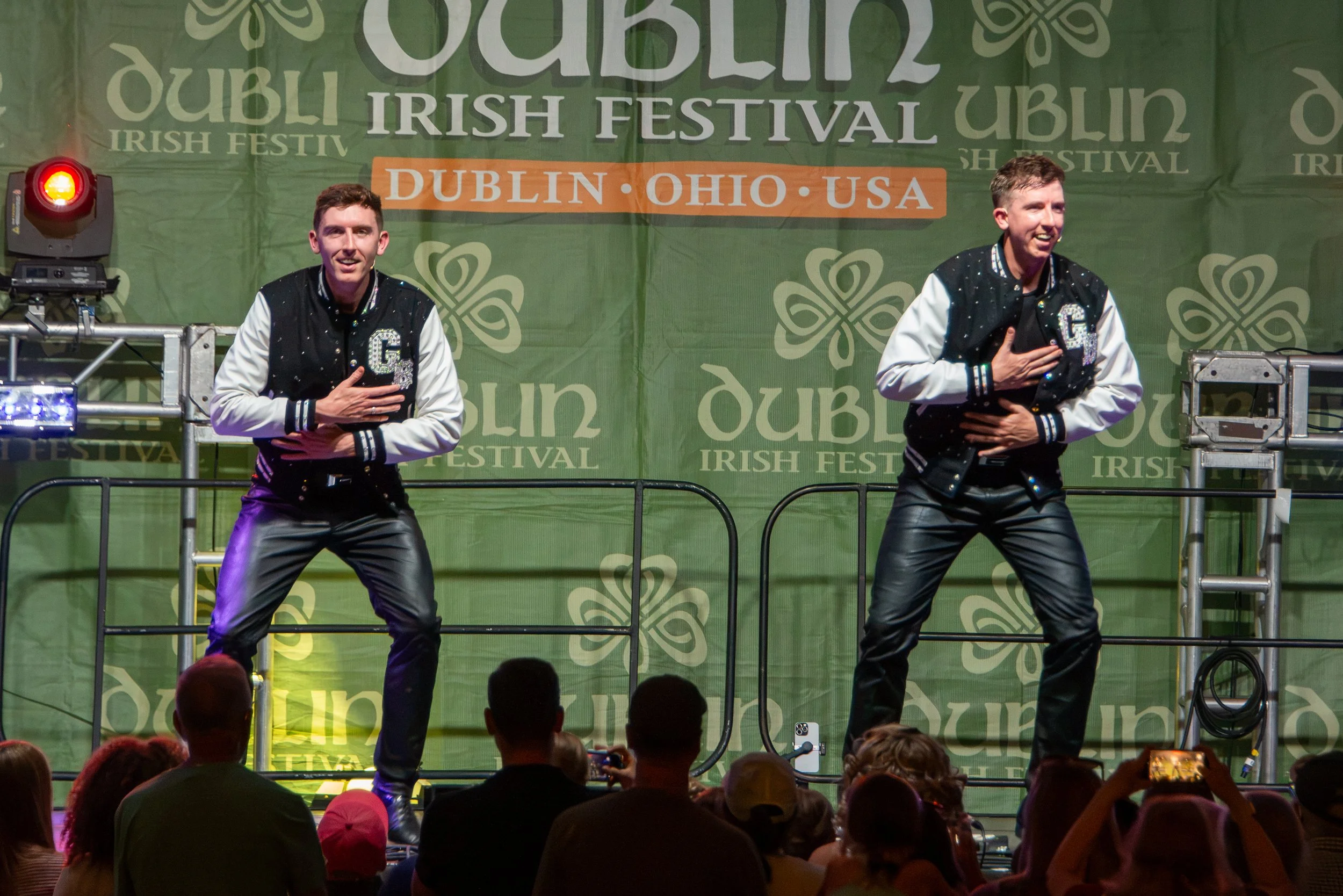 Two male performers dancing on stage at an Irish festival in Dublin, Ohio, USA. They are wearing black and white jackets with a letter 'G' on the chest, and have their right hands over their hearts. The background features a large green banner with s