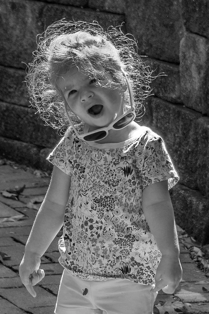 A young girl with wild curly hair, wearing sunglasses around her neck and a floral shirt, standing outdoors on a stone path next to a brick wall, smiling or shouting.