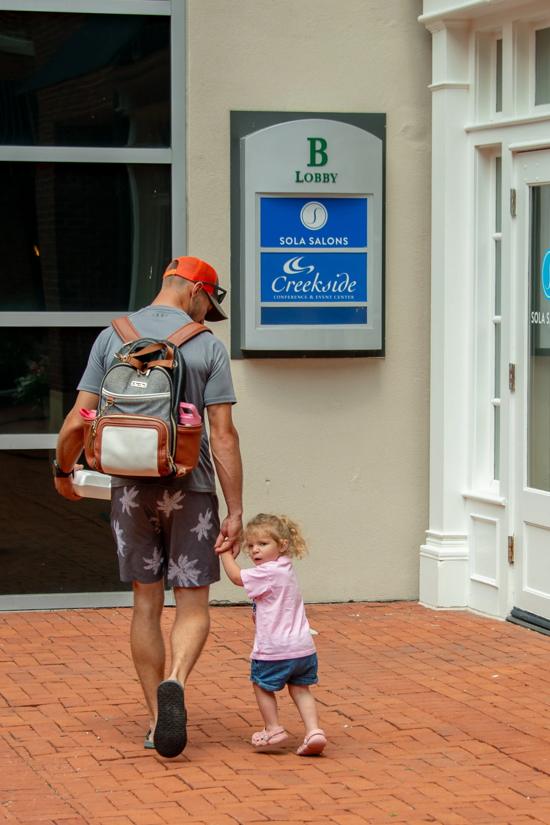A man with a backpack and a young girl holding hands walking on a brick sidewalk in front of a building with a sign for Sola Salons, Creekside conference and event center, and the building entrance with white trim.