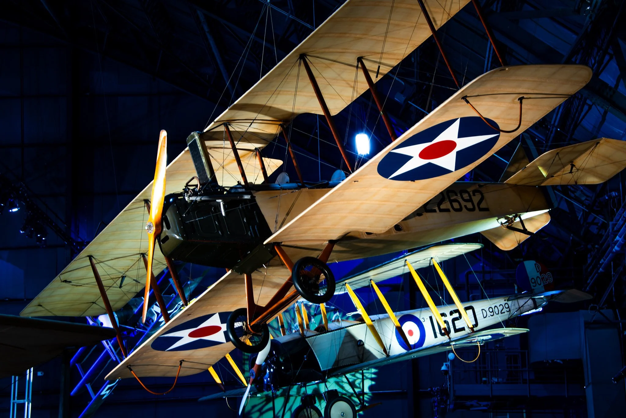 Multiple vintage biplanes with wood wings and navy blue fuselages are displayed in an indoor exhibition, illuminated with blue lighting.