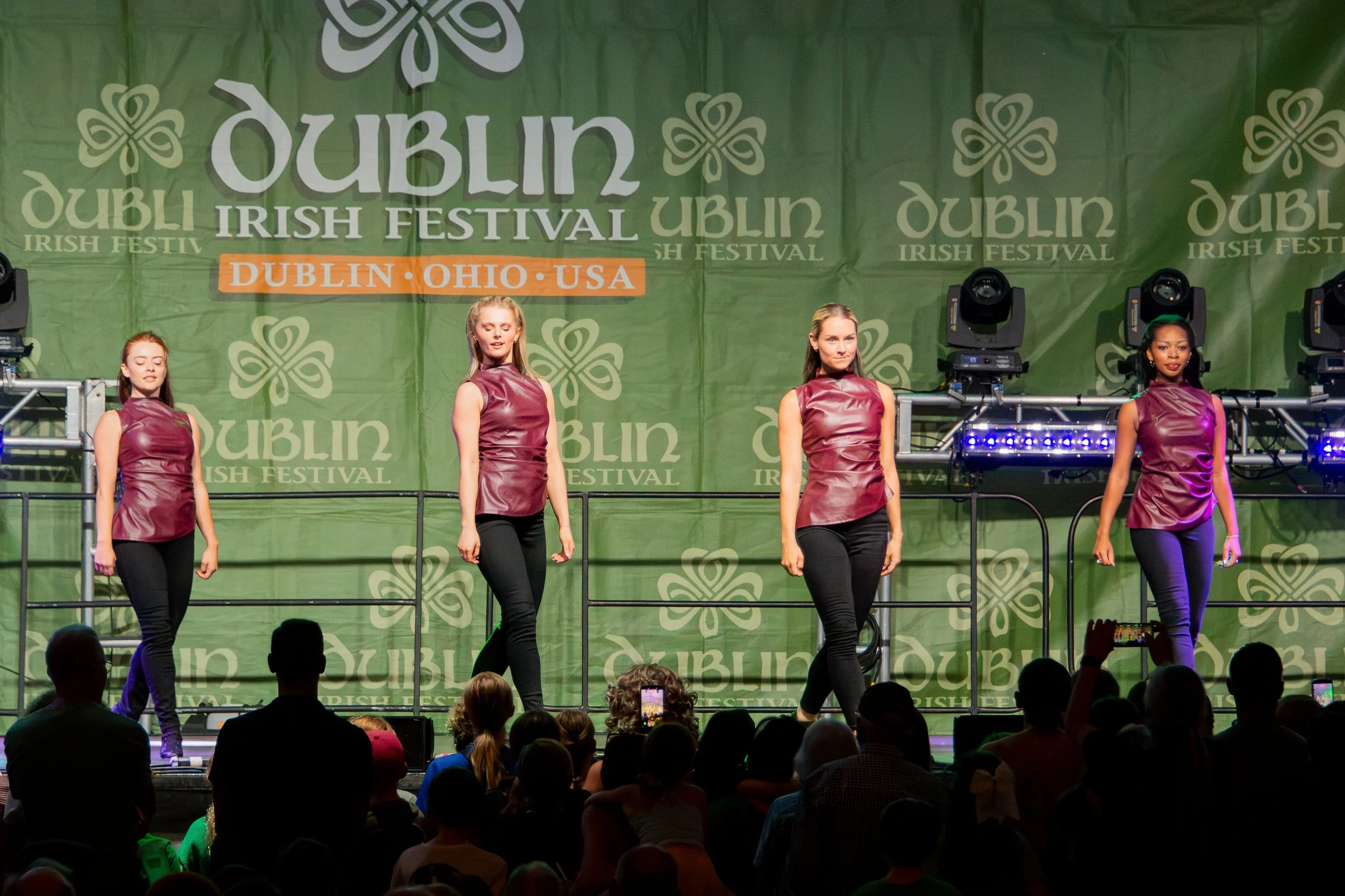 Four women in maroon tops and black pants performing a dance on stage at the Dublin Irish Festival, with a green backdrop decorated with shamrock designs and the festival name.