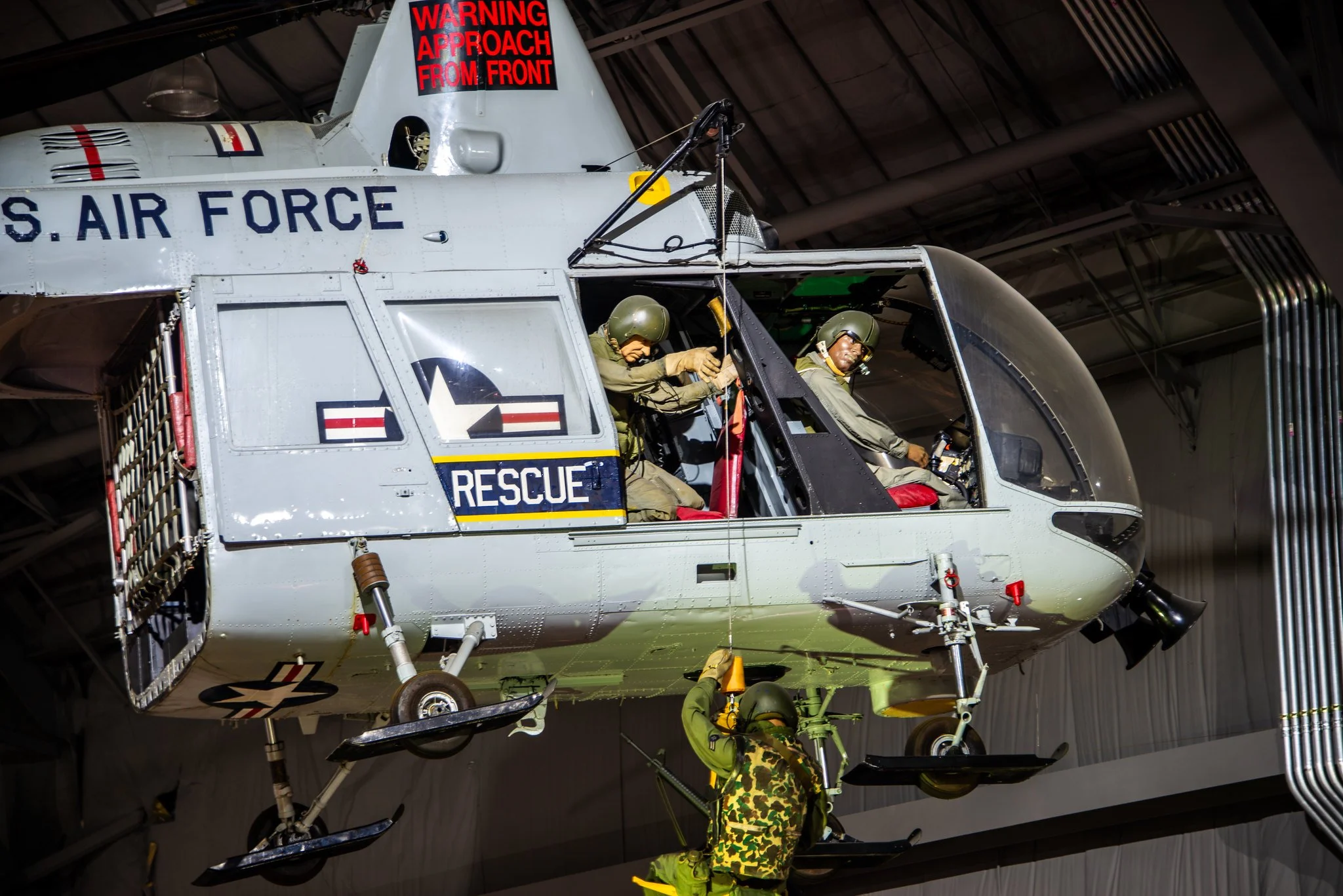 Military helicopter inside a hangar with personnel in helmets working on it. A red warning sign on the helicopter says, 'Warning Approach From Front.'