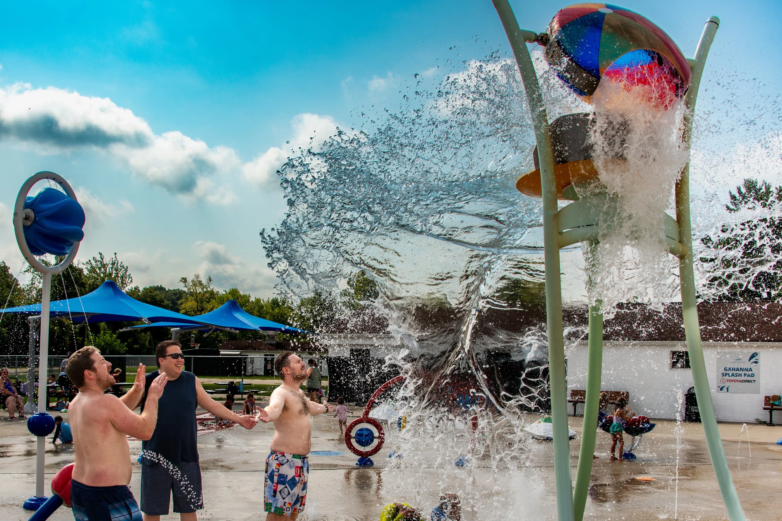 People enjoying water spray from a splash pad on a sunny day, with children and adults playing and laughing.