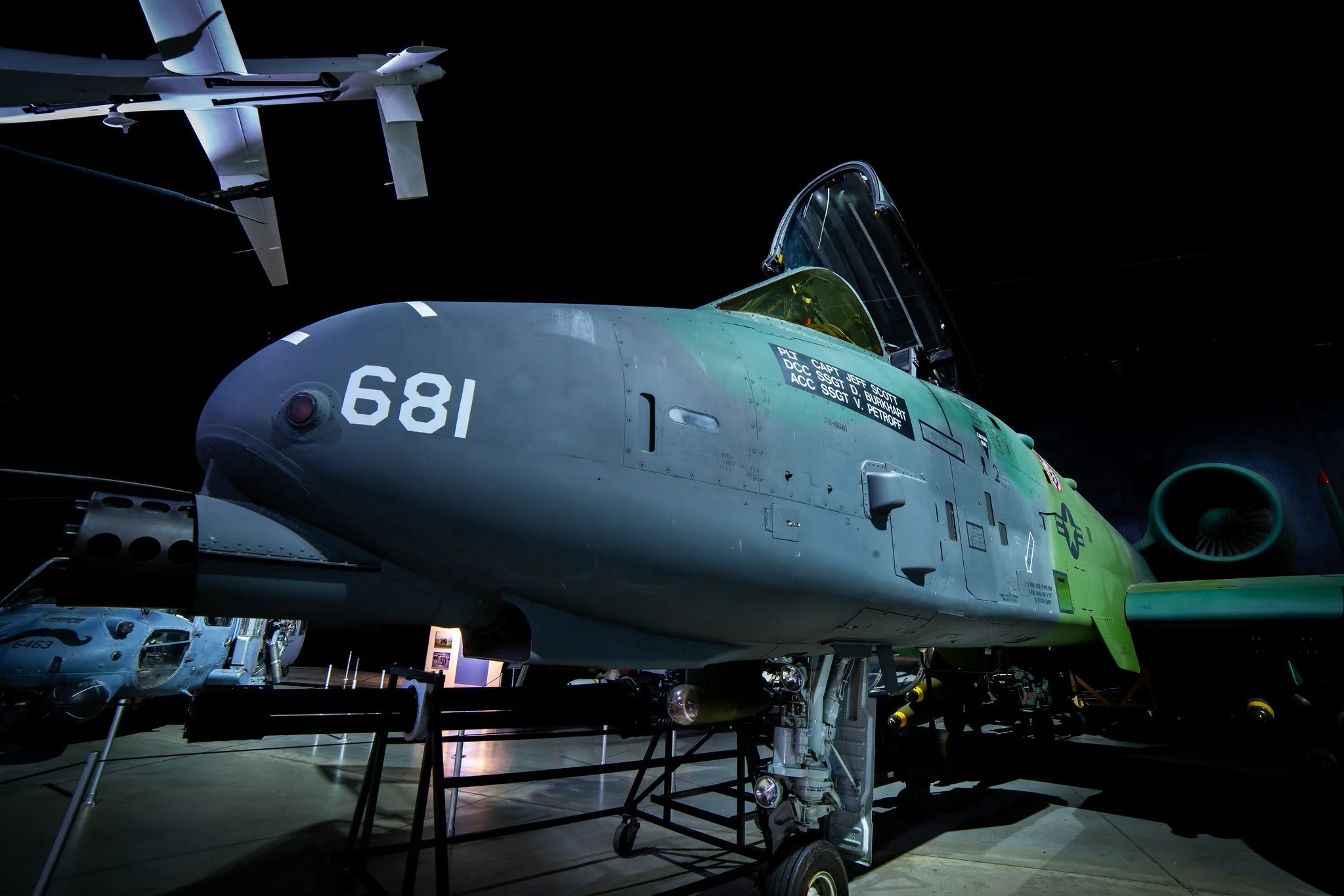 Two fighter jets displayed indoors, one in front with the cockpit open and the other in the background. The jets are gray with various markings and labels, under bright lighting with a dark background.