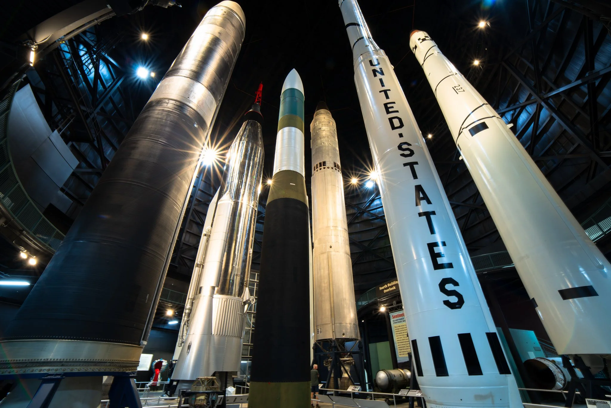 Various rockets on display inside a museum with dim lighting and blue accents, including a large metallic rocket and a white rocket with black markings.