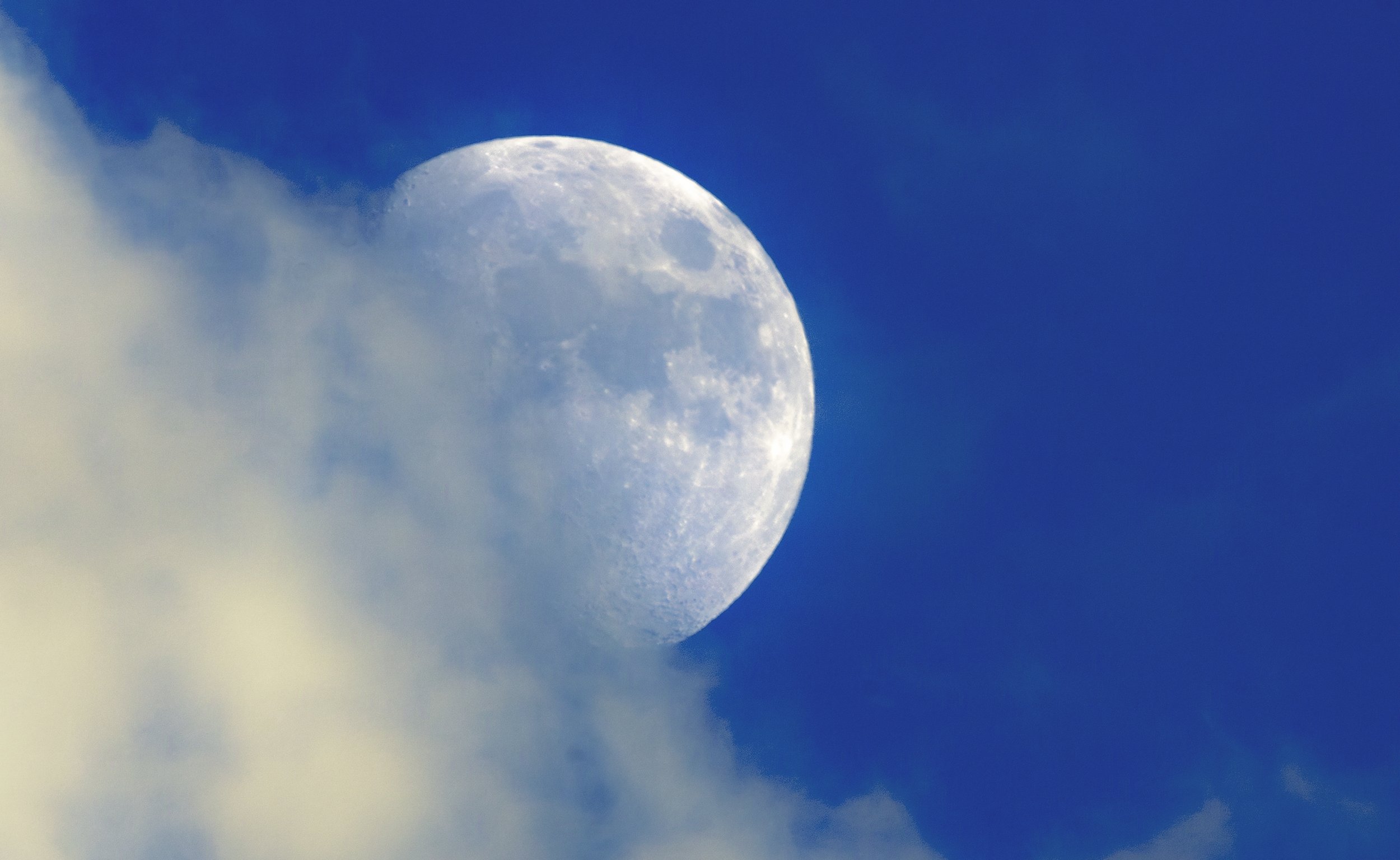Close-up of the Moon in the daytime sky with a blue background and clouds partially covering it.