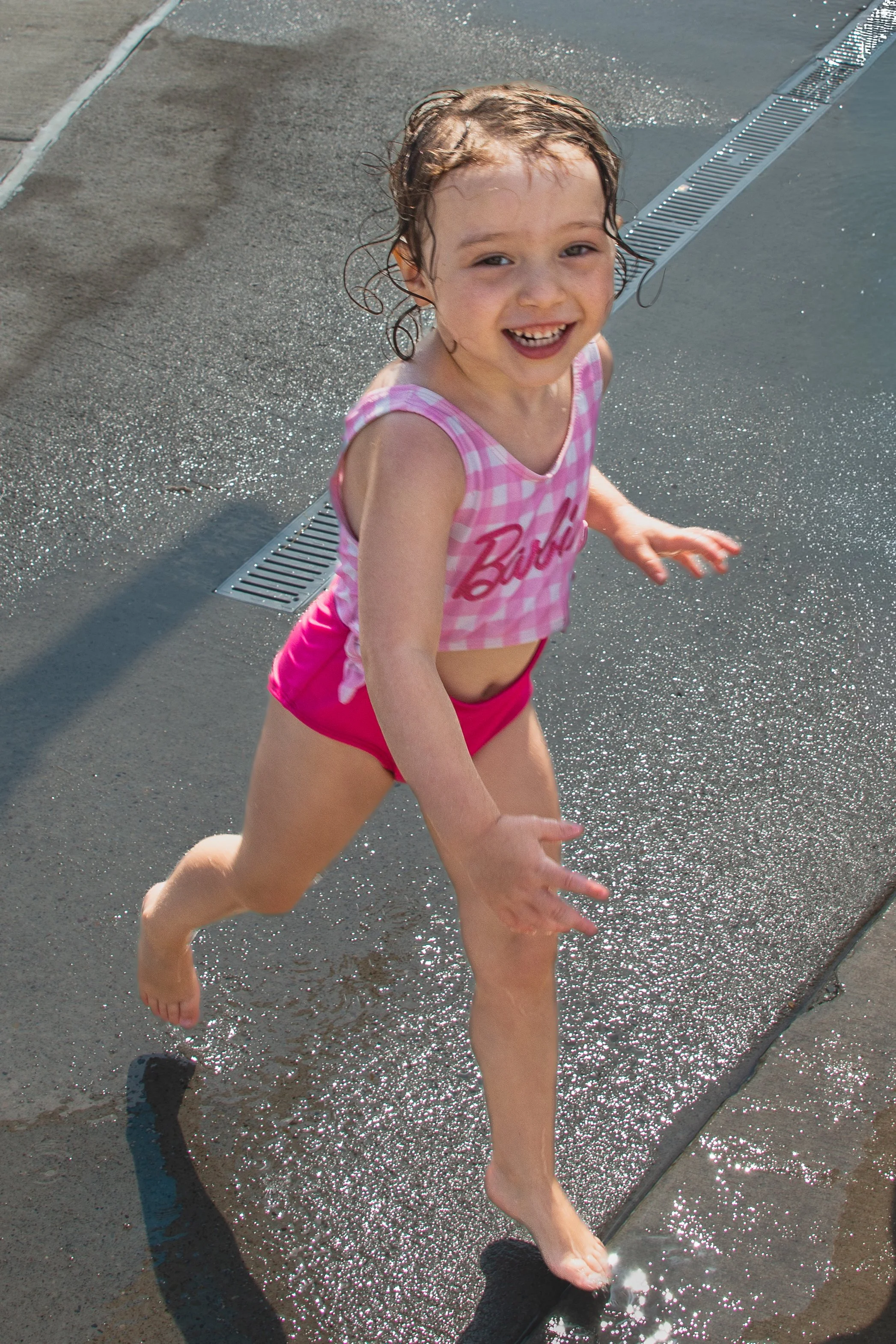 Young girl with wet hair and pink checkered shirt playing and running barefoot near water drainage on wet concrete sidewalk.