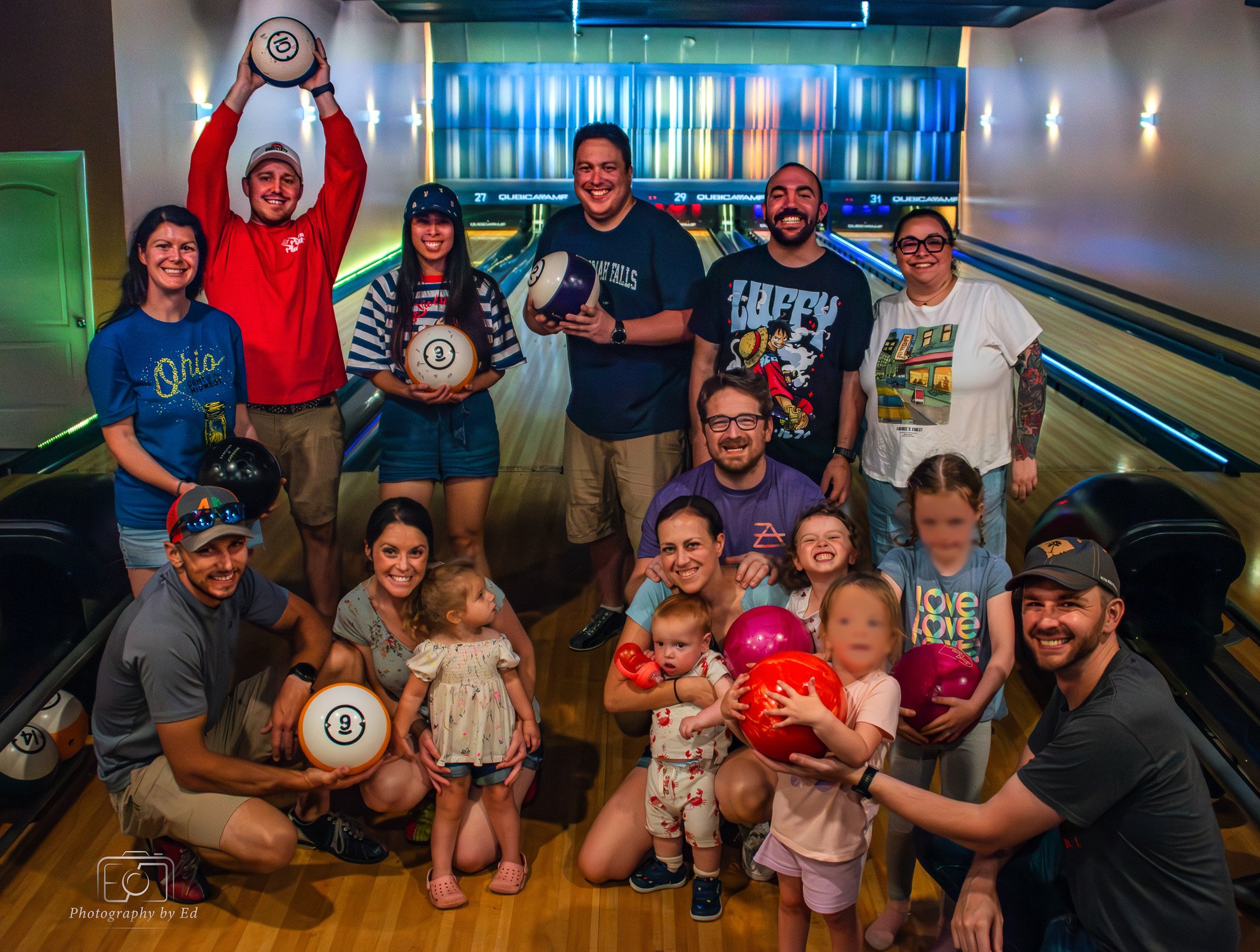 A group of people at a bowling alley, smiling and holding bowling balls. The group includes children and adults, with some children holding balloons. The background shows bowling lanes with digital score displays and colorful lighting. The atmosphere appears fun and lively.