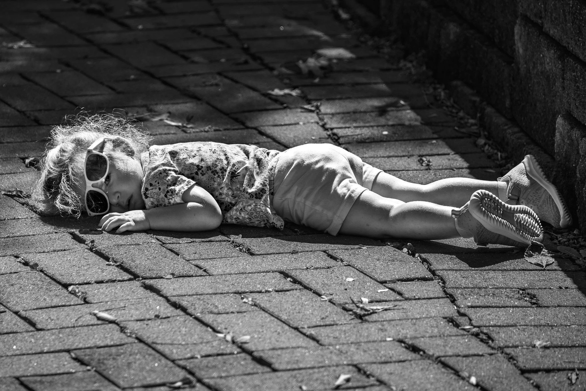 A young girl lying on a brick sidewalk next to a stone wall, wearing sunglasses, a floral shirt, and shorts, with her eyes closed.