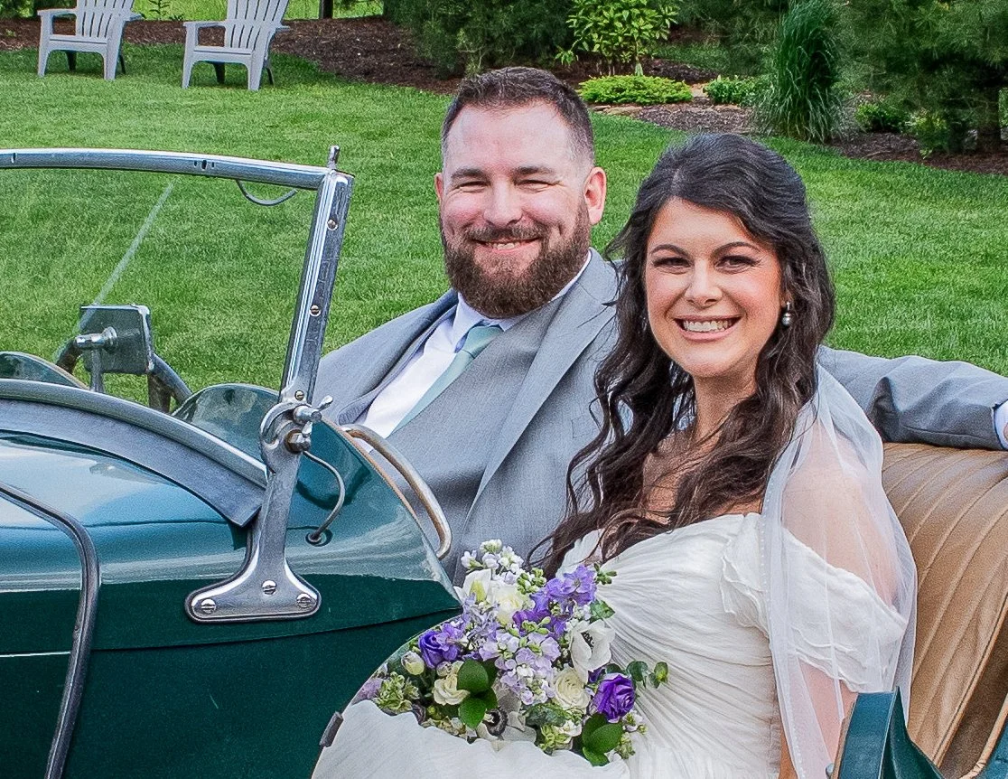 A newlywed couple sitting in a vintage car outdoors, with the bride holding a bouquet of flowers and smiling at the camera.