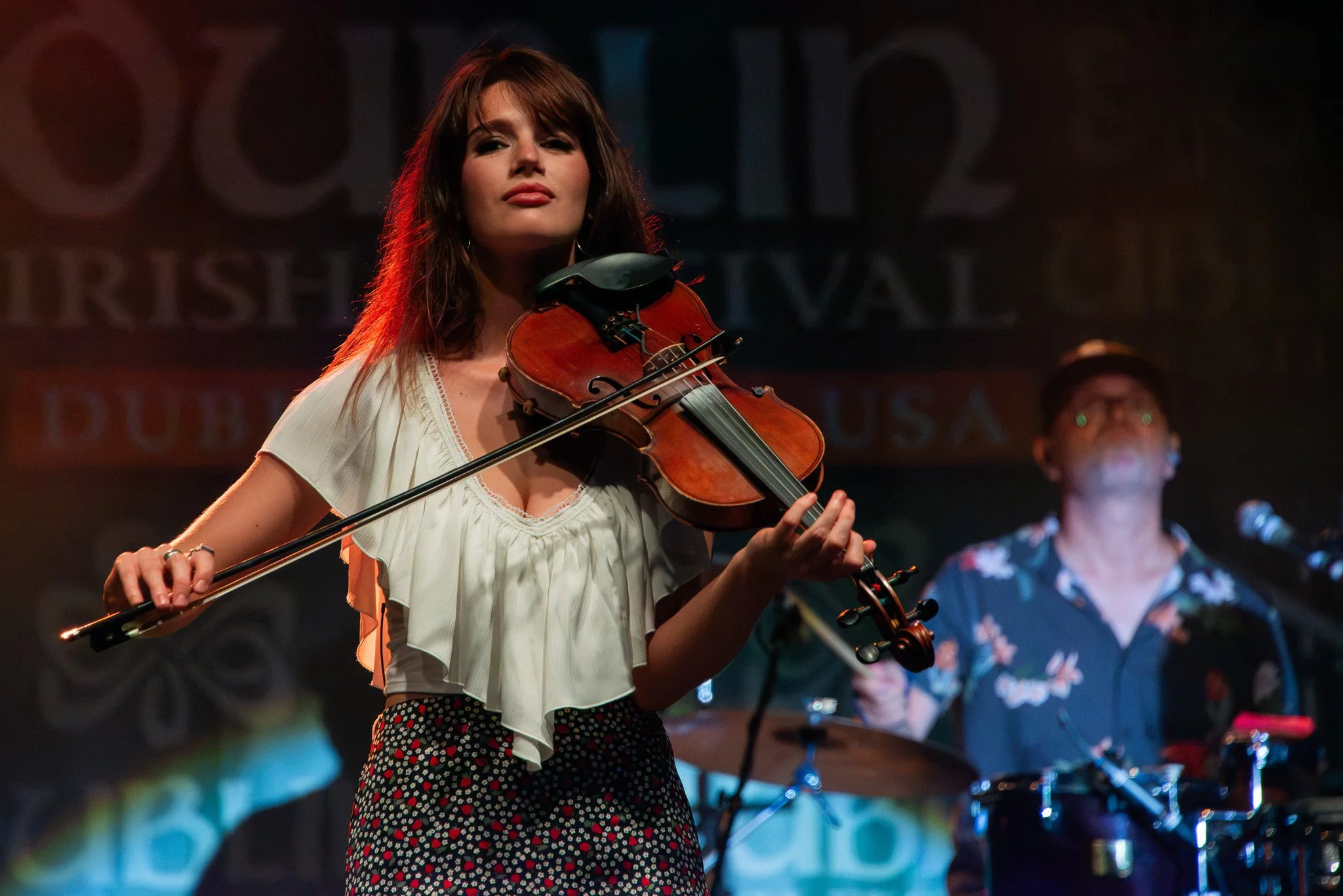 Natalya Kay playing the violin on stage, wearing a white blouse and patterned pants, with Ryan Lacey in floral shirt playing drums in the background at the Dublin Irish Festival.