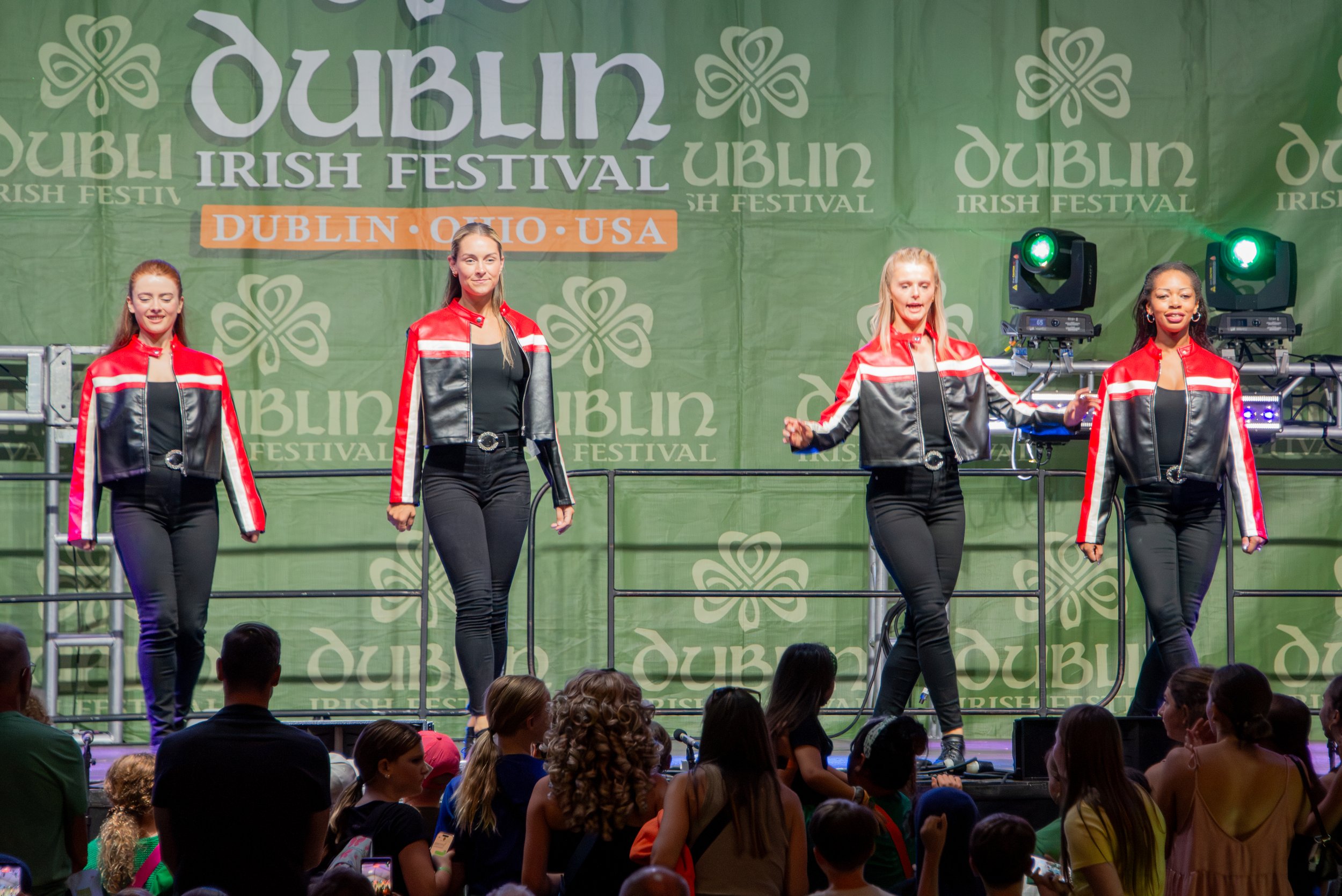 Four women dressed in red and black jackets performing on stage at the Irish Festival in Dublin, Ohio, with a green backdrop and an audience watching.
