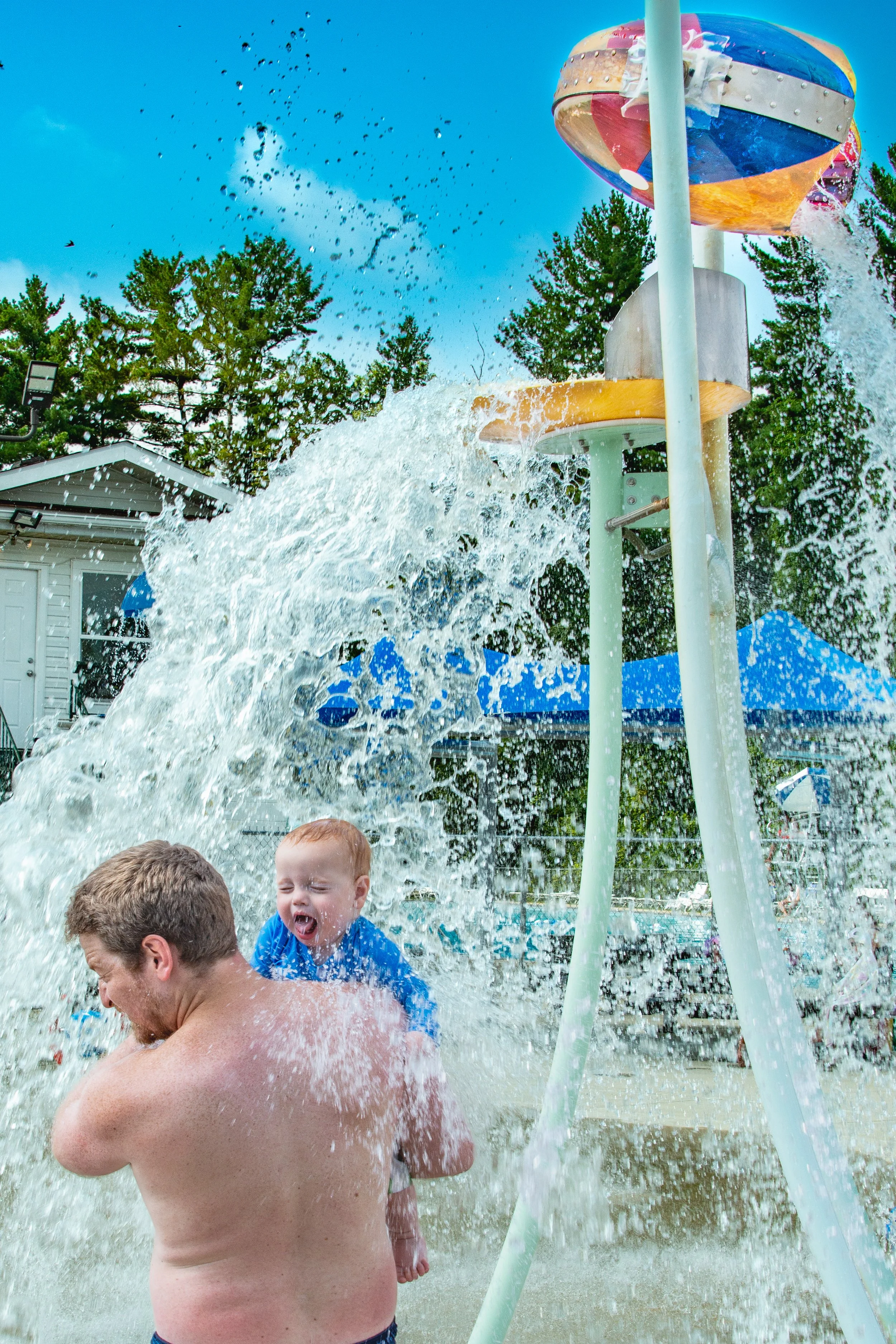 Children and adults playing under a water splash feature at a splash pad, with water spraying from a large colorful water tower, trees in the background, and a bright blue sky.