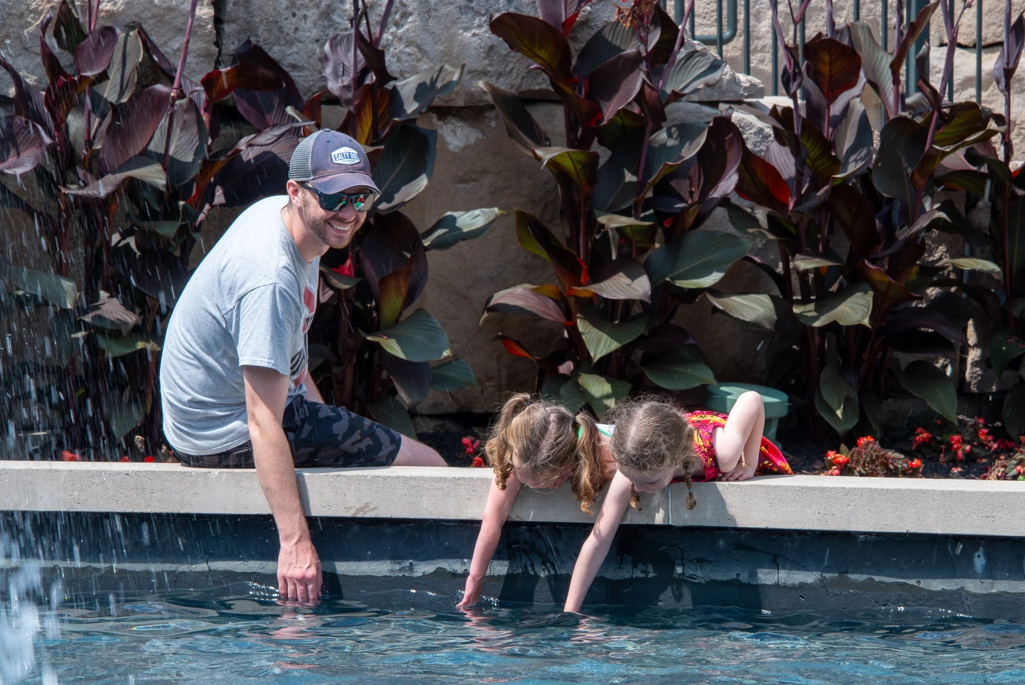 A man wearing sunglasses, a hat, and a t-shirt is crouching by a water feature, smiling and helping two children submerged in the water to touch it. Behind them are large purple and green plants and a stone wall.