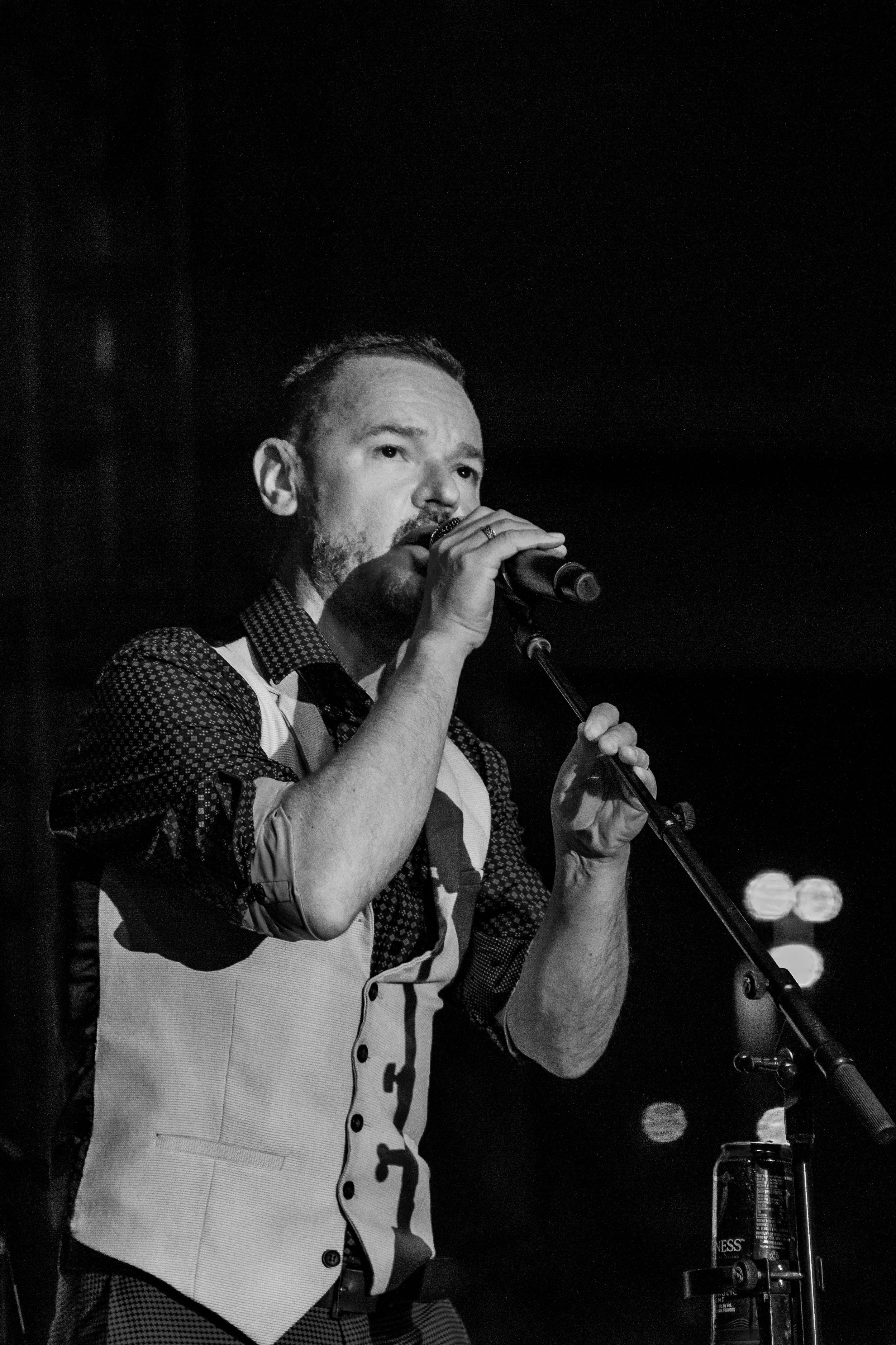 Black and white photograph of a Patrick Murphy of Gaelic Storm singing into a microphone on stage, wearing a patterned shirt and a vest, with stage lights in the background.