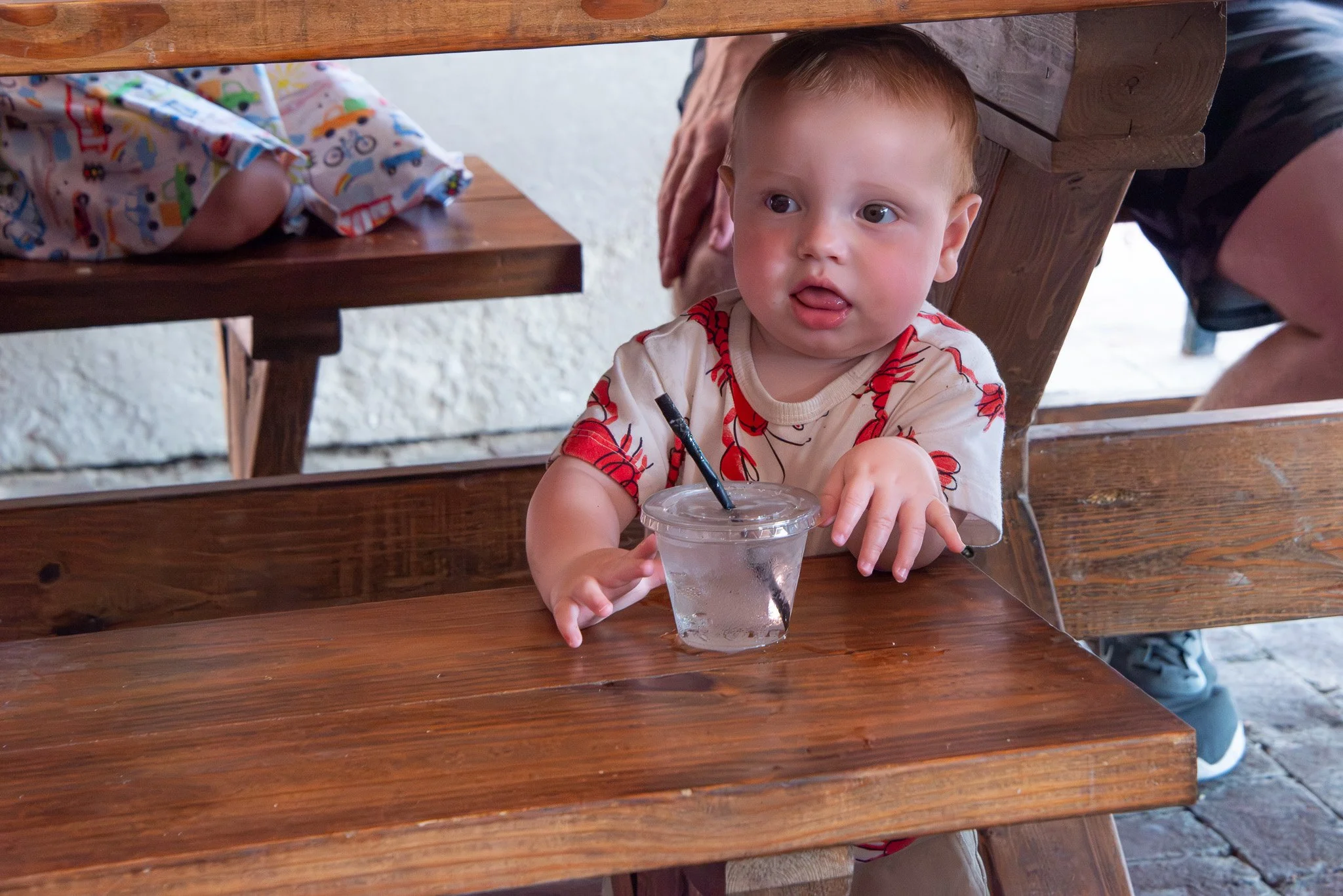 A young boy sitting at a wooden table, holding a clear plastic cup with a straw, sticking out his tongue slightly, with a curious expression, and wearing a light-colored t-shirt with red designs. There is a person next to him, partially visible, and 