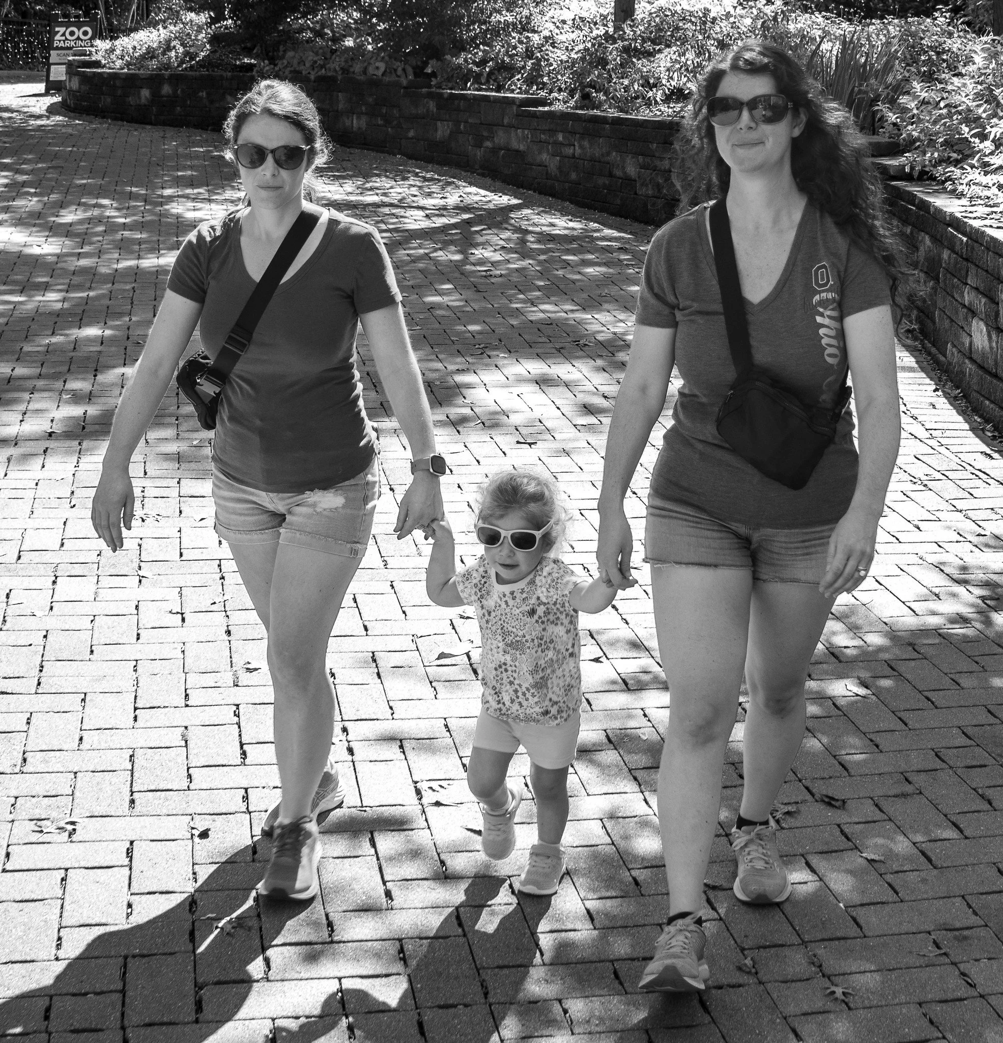 Two women and a small child walking outdoors in sunlight with shadows cast on brick pavement, foliage, and a brick wall in the background, all wearing sunglasses and casual clothing.