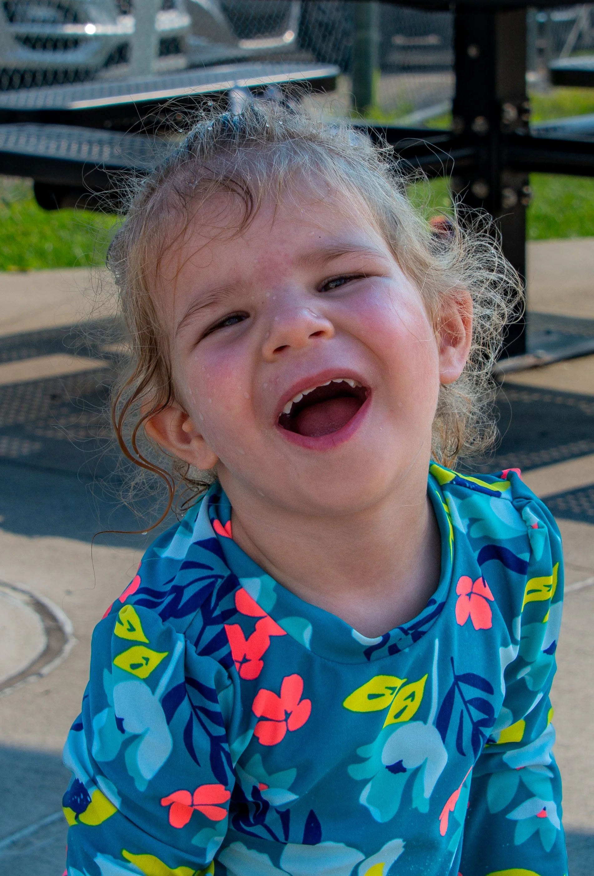 Close-up of a young girl with curly blonde hair, wearing a colorful, blue floral-patterned swimsuit, smiling widely with her mouth open, at a playground with metal climbing structures in the background.