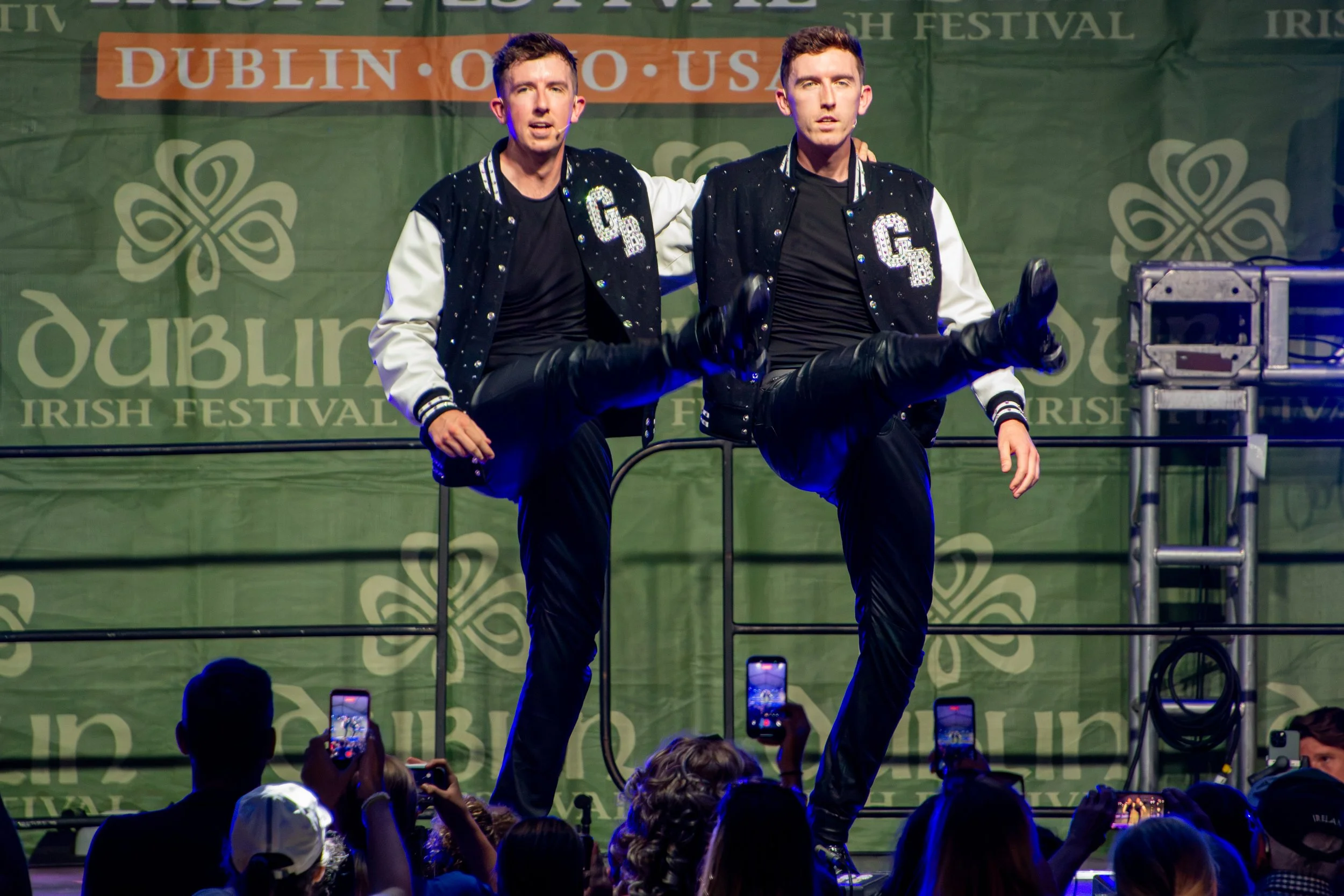 The Gardiner Brothers are performing on a stage at the Dublin Irish Festival holding a leg lift pose, wearing matching black and white jackets with rhinestones, black shirts, and black pants, with an audience holding up phones to record the performan