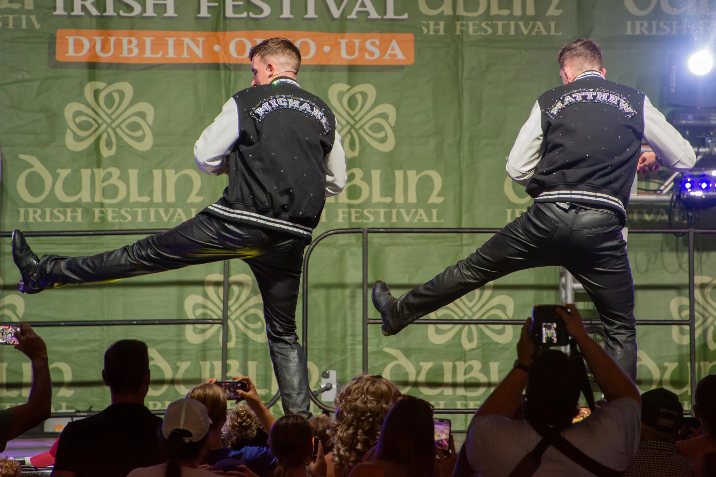 Two male dancers wearing black and white jackets with 'Michael' and 'Matthew' on the back, performing on stage at the Irish Fest in Dublin, Ohio, with a green background banner and audience members taking photos.