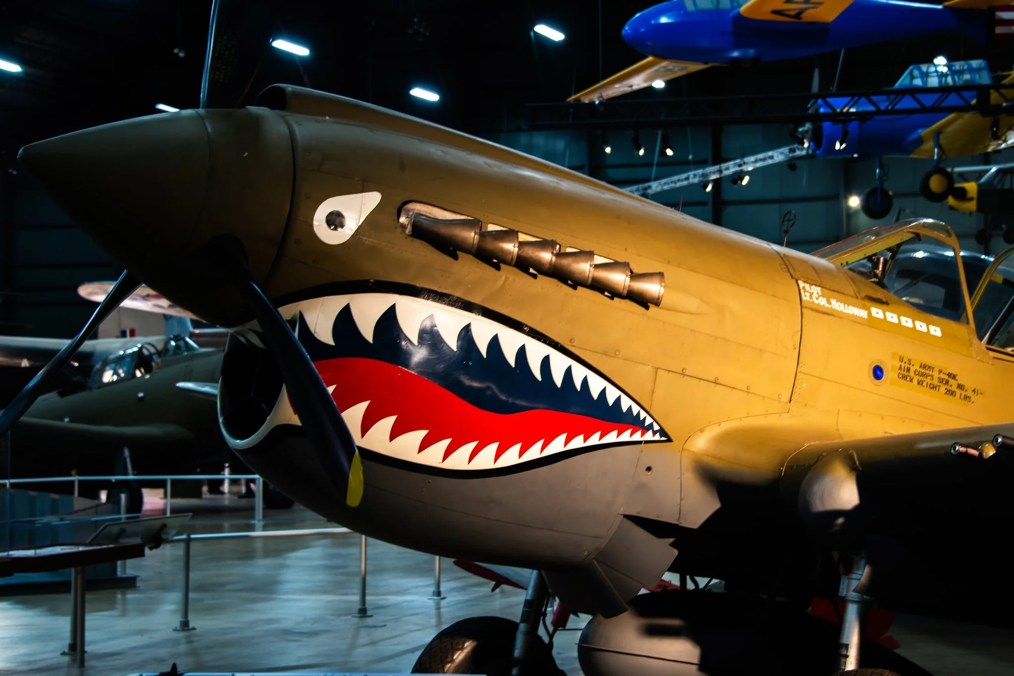 A vintage fighter plane with shark face artwork on its nose, displayed in an aviation museum with other aircrafts visible in the background.