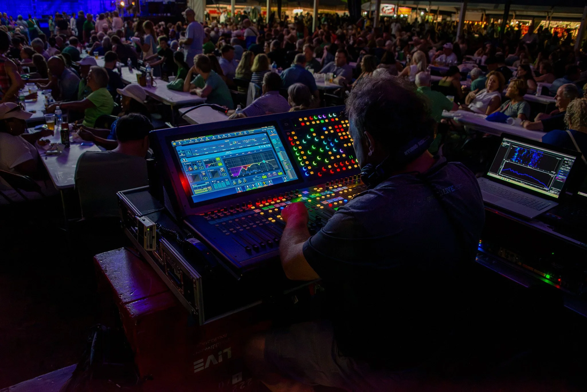 A crowd of people seated at tables in a large, open-air venue during nighttime. In the foreground, a person is operating DJ equipment with colorful lights and computer screens.