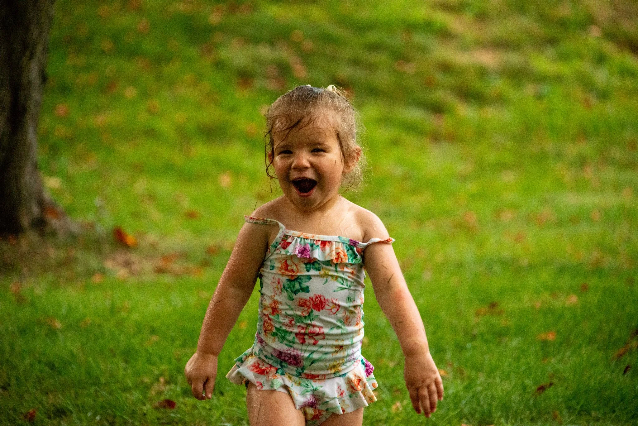 A young girl with wet hair wearing a colorful floral dress, standing on green grass with trees in the background, smiling with her mouth open.
