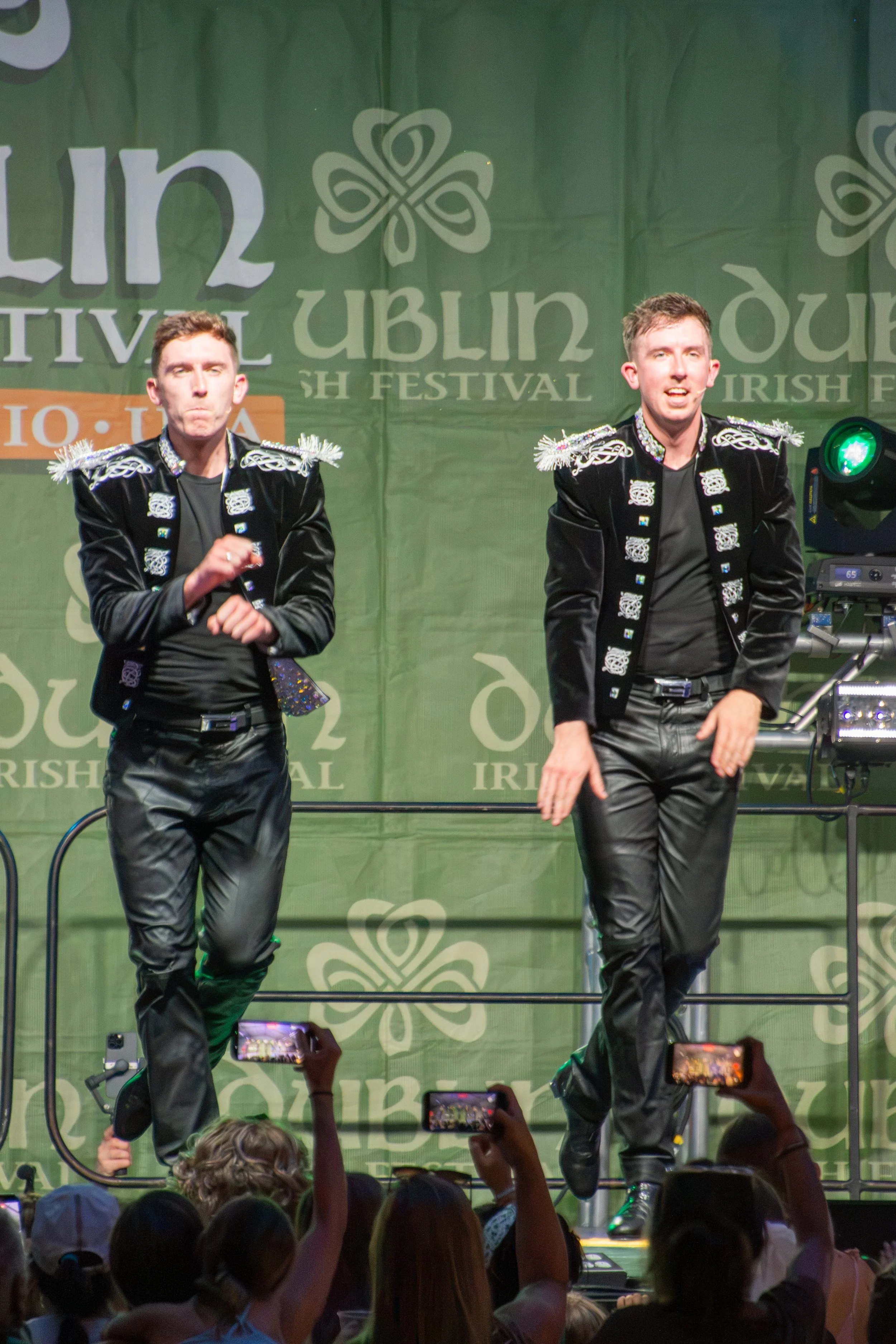 Two performers on stage dressed in black costumes with silver decorative elements, performing at an Irish festival. Audience members are taking photos with their phones.